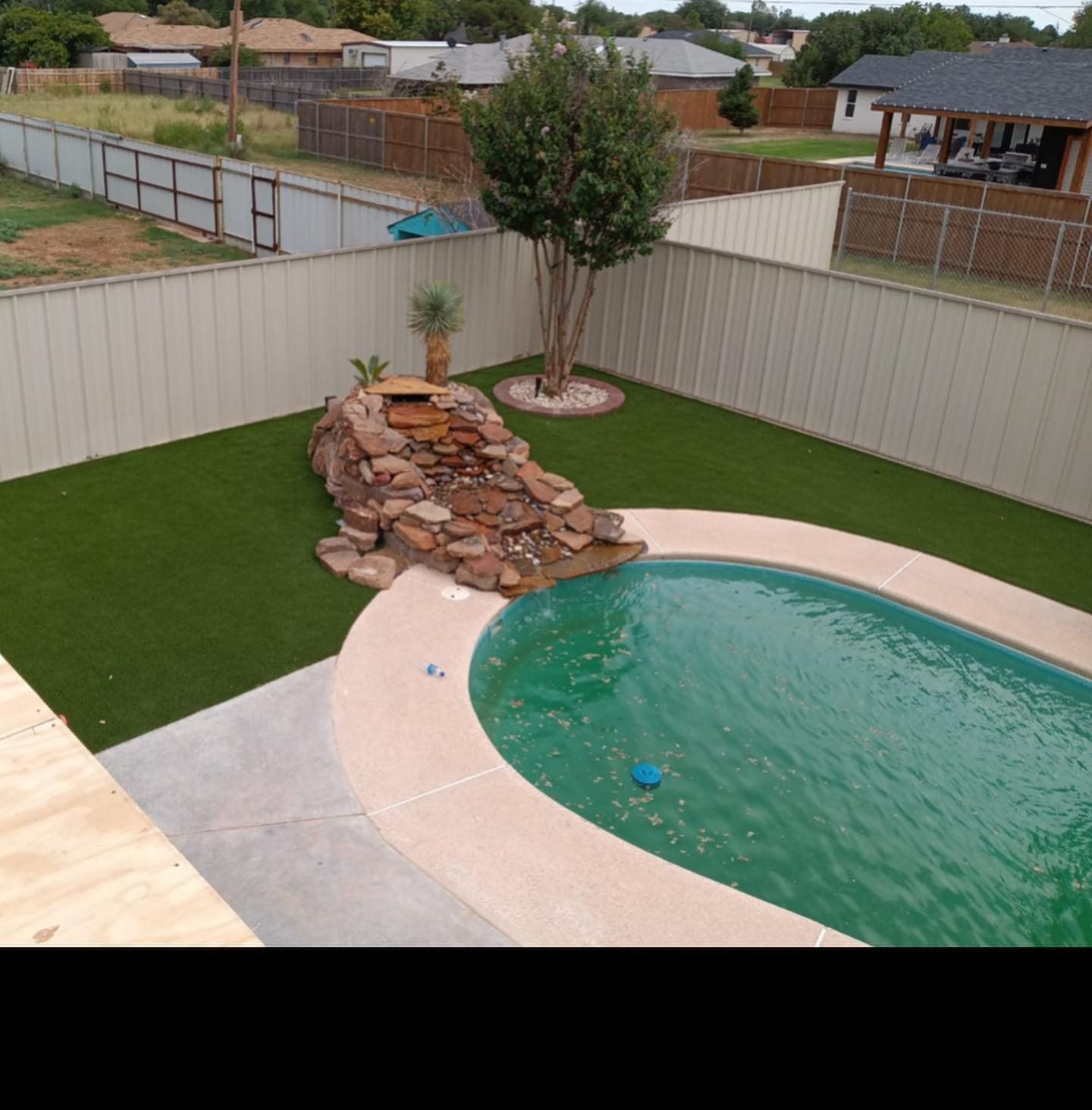 Backyard pool with rock waterfall, green lawn, and beige privacy fence