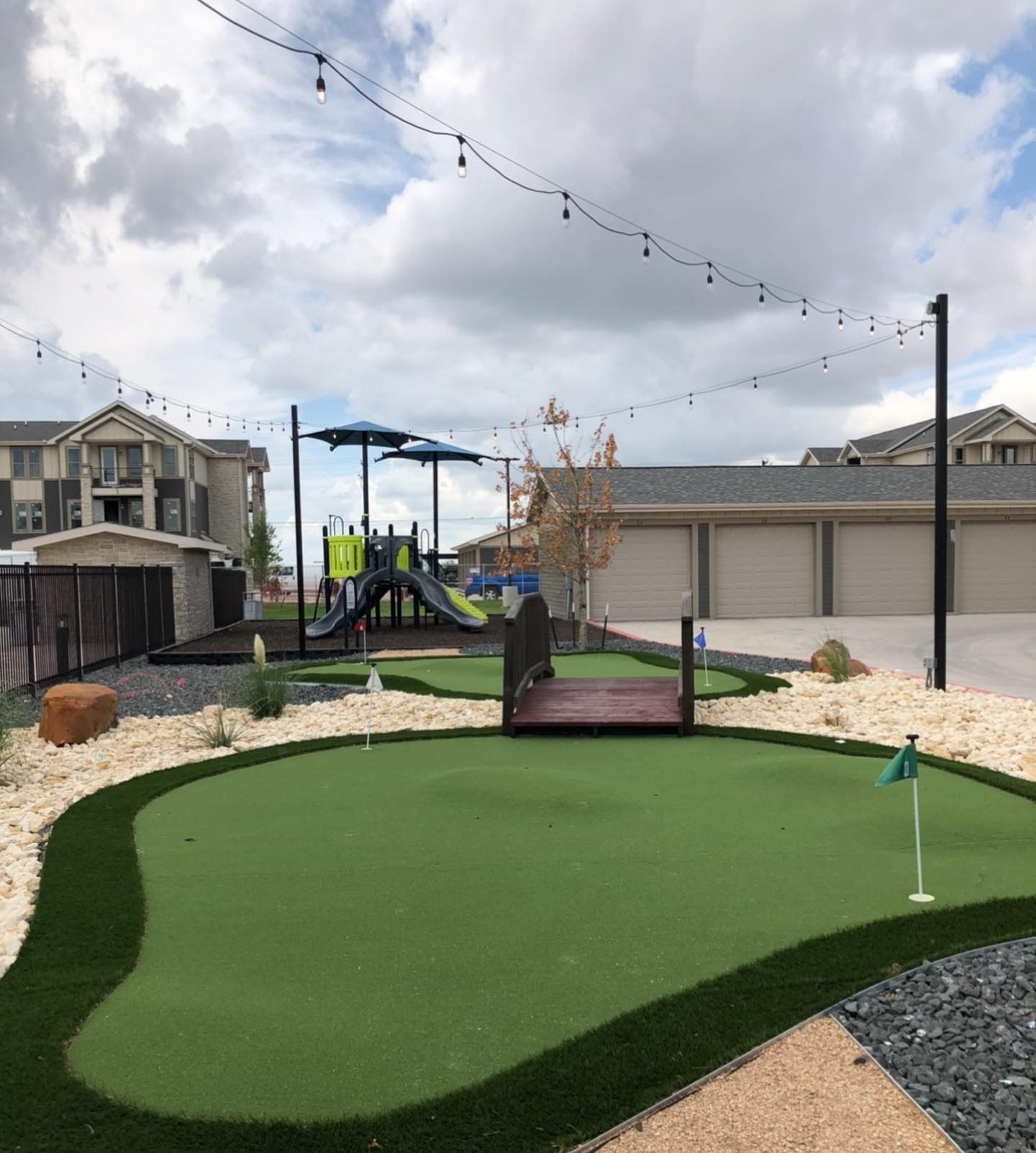 Backyard mini golf green with a small bridge, string lights, and playground in the background