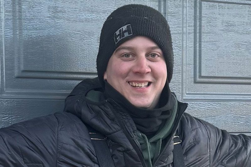 Man in black beanie, coat, and neck gaiter smiles in front of a blue garage door.