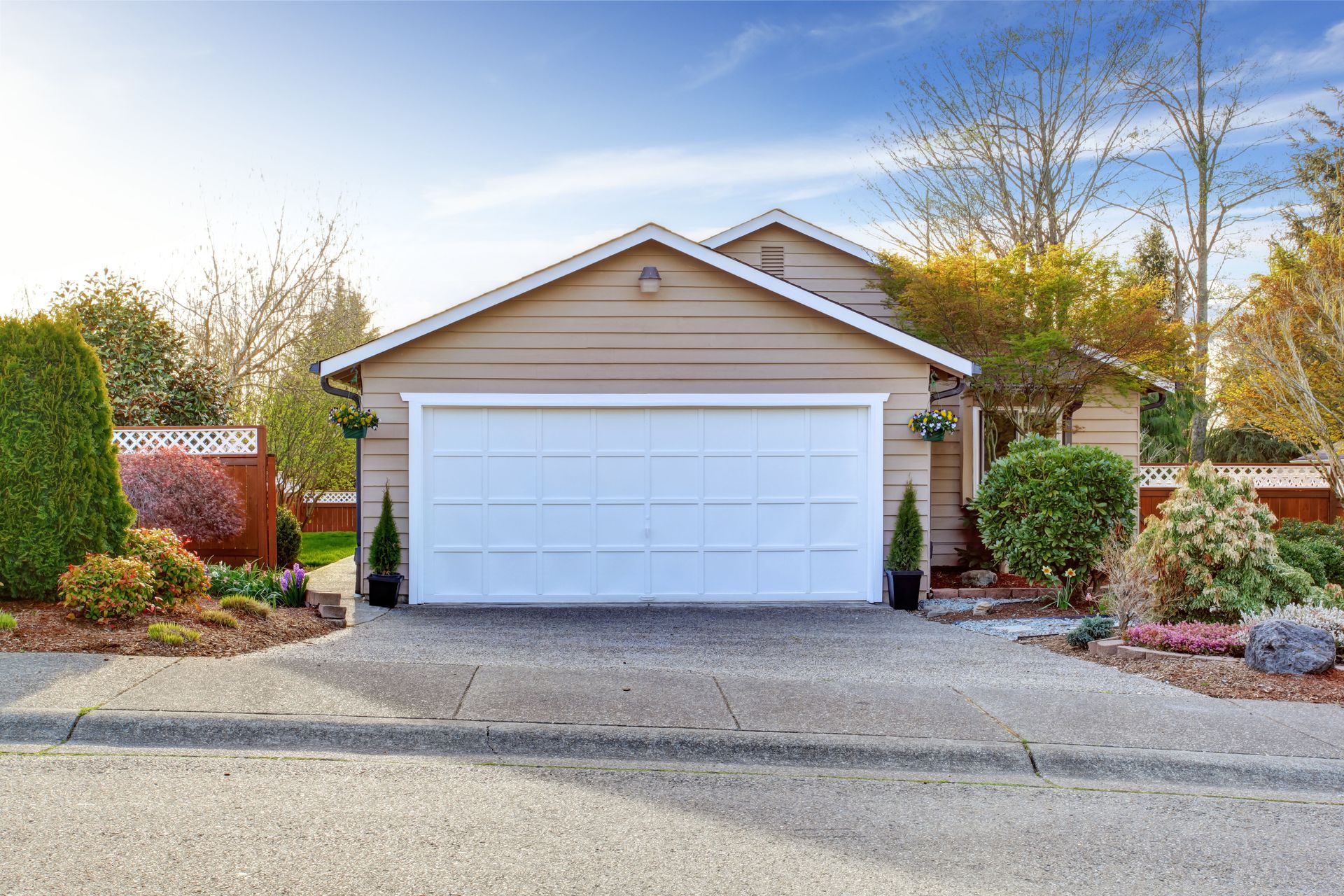 Garage with white door, tan siding, and concrete driveway, surrounded by landscaping under a blue sky.