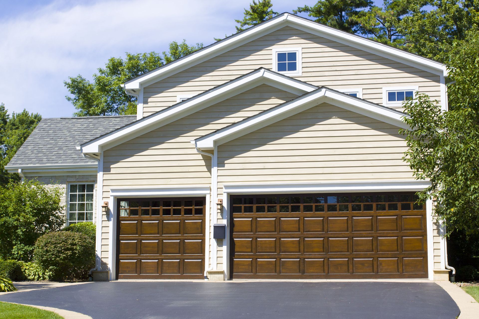 Tan two-story house with brown garage doors, driveway, and surrounding greenery under a blue sky.