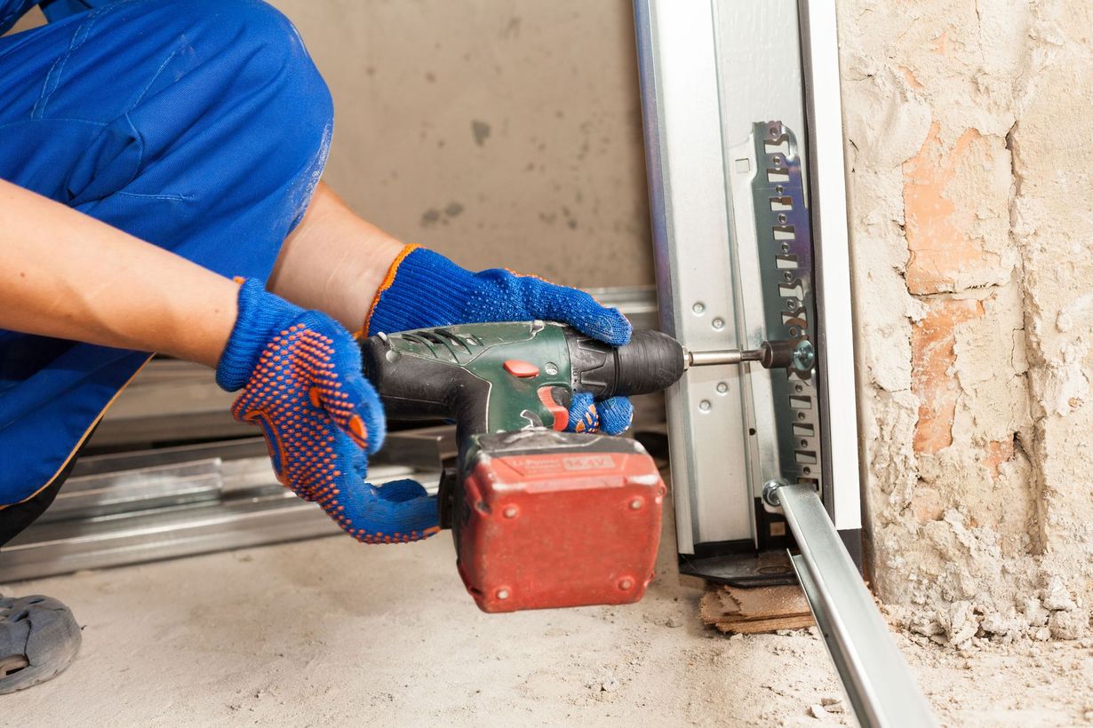 Person in blue coveralls using a drill on a garage door track; brick wall in the background.