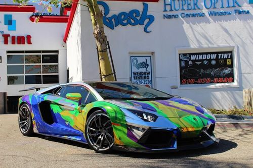 Lamborghini with colorful wrap parked in front of a "Fresh Tint" shop, sunny day.