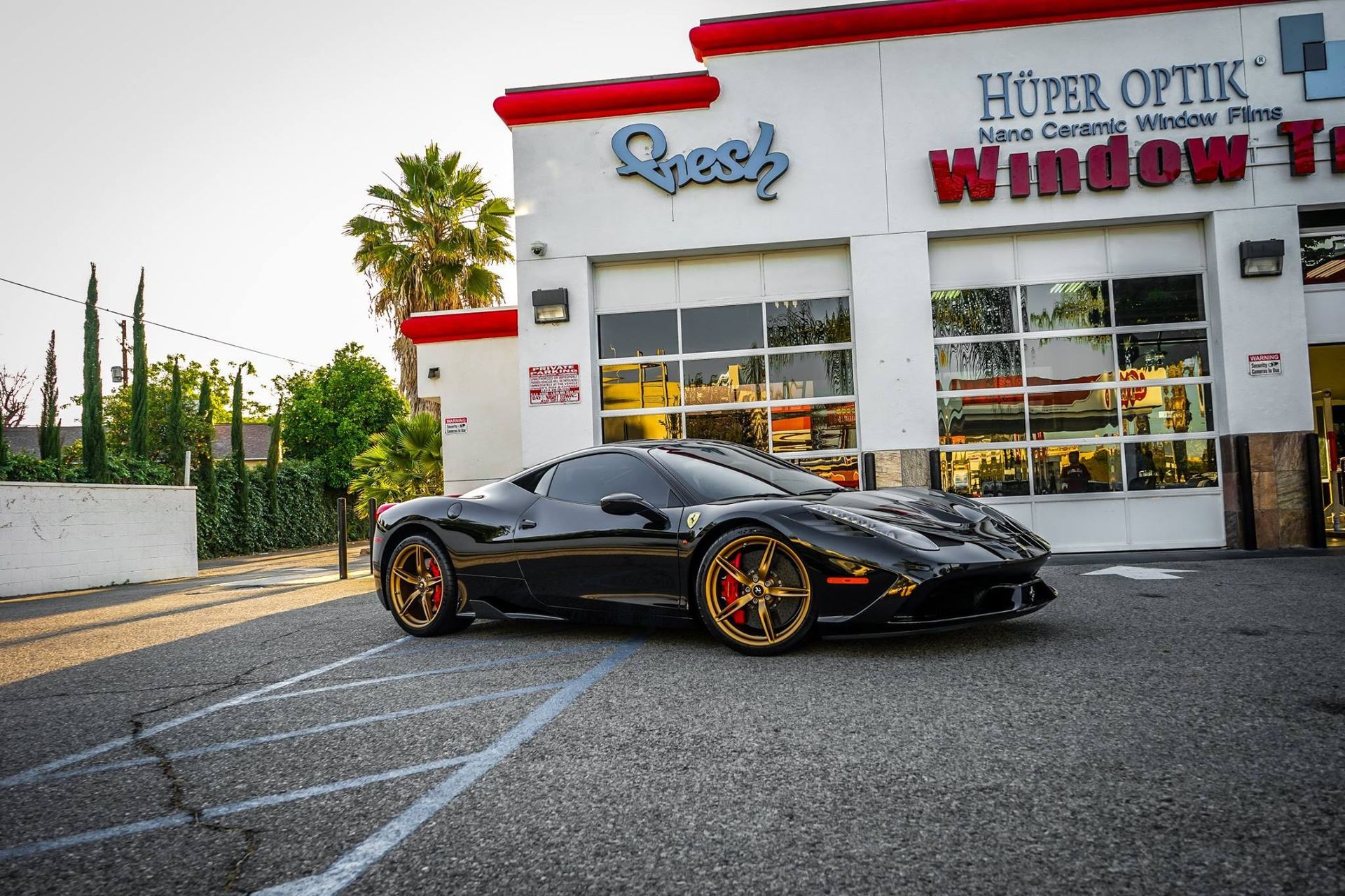 Black sports car parked in front of a window tint shop; gold and red wheels.