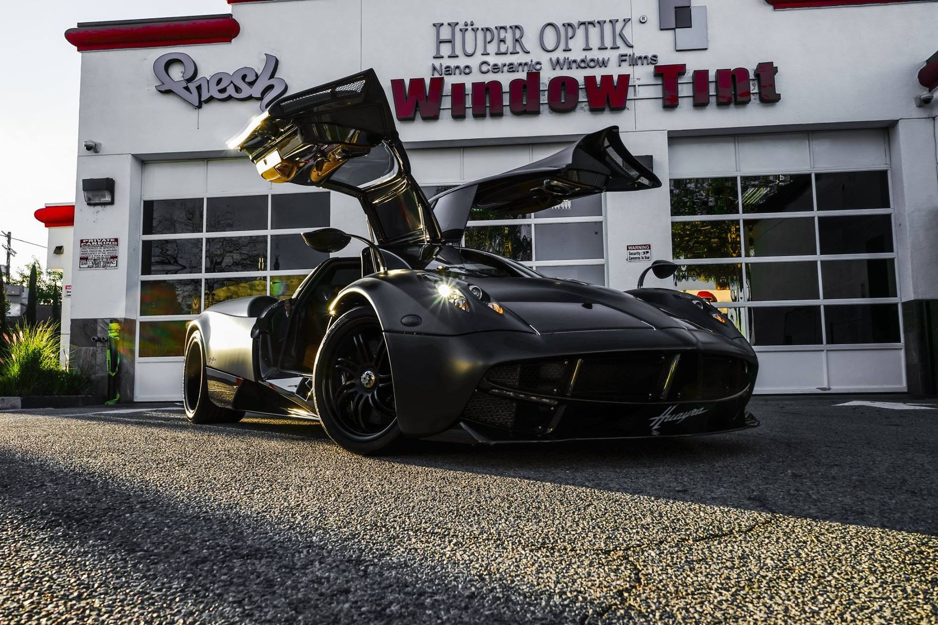 Black Pagani Huayra with gullwing doors open in front of a window tint shop.