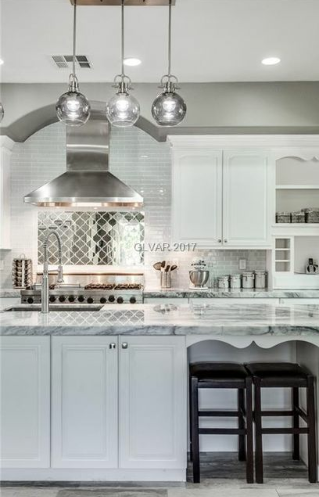 Elegant white kitchen with stainless steel appliances, marble countertops, and dark bar stools. Three pendant lights hang above.