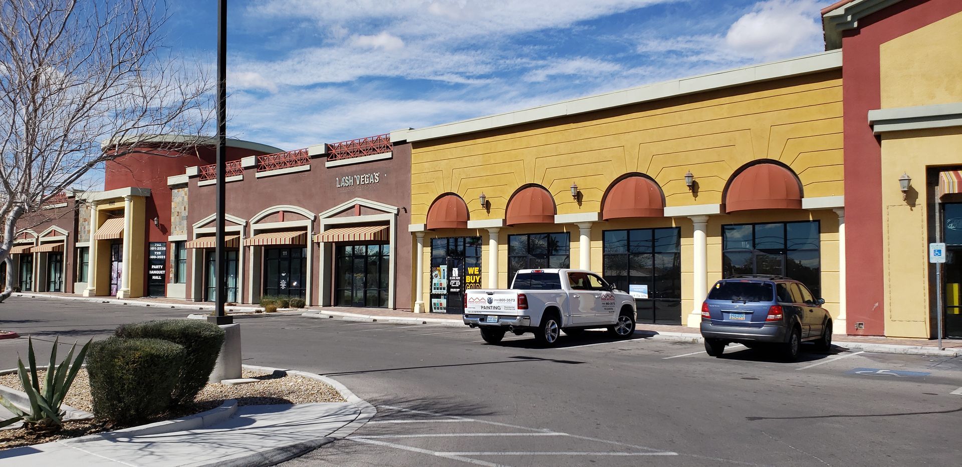 A commercial building with a row of shops. A white pickup truck and a grey car are parked in front. Blue sky with clouds overhead.