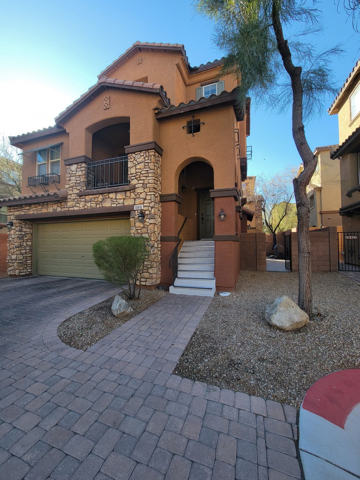 Three-story stucco home with stone accents and a small front yard with a brick driveway. A green garage door is visible.