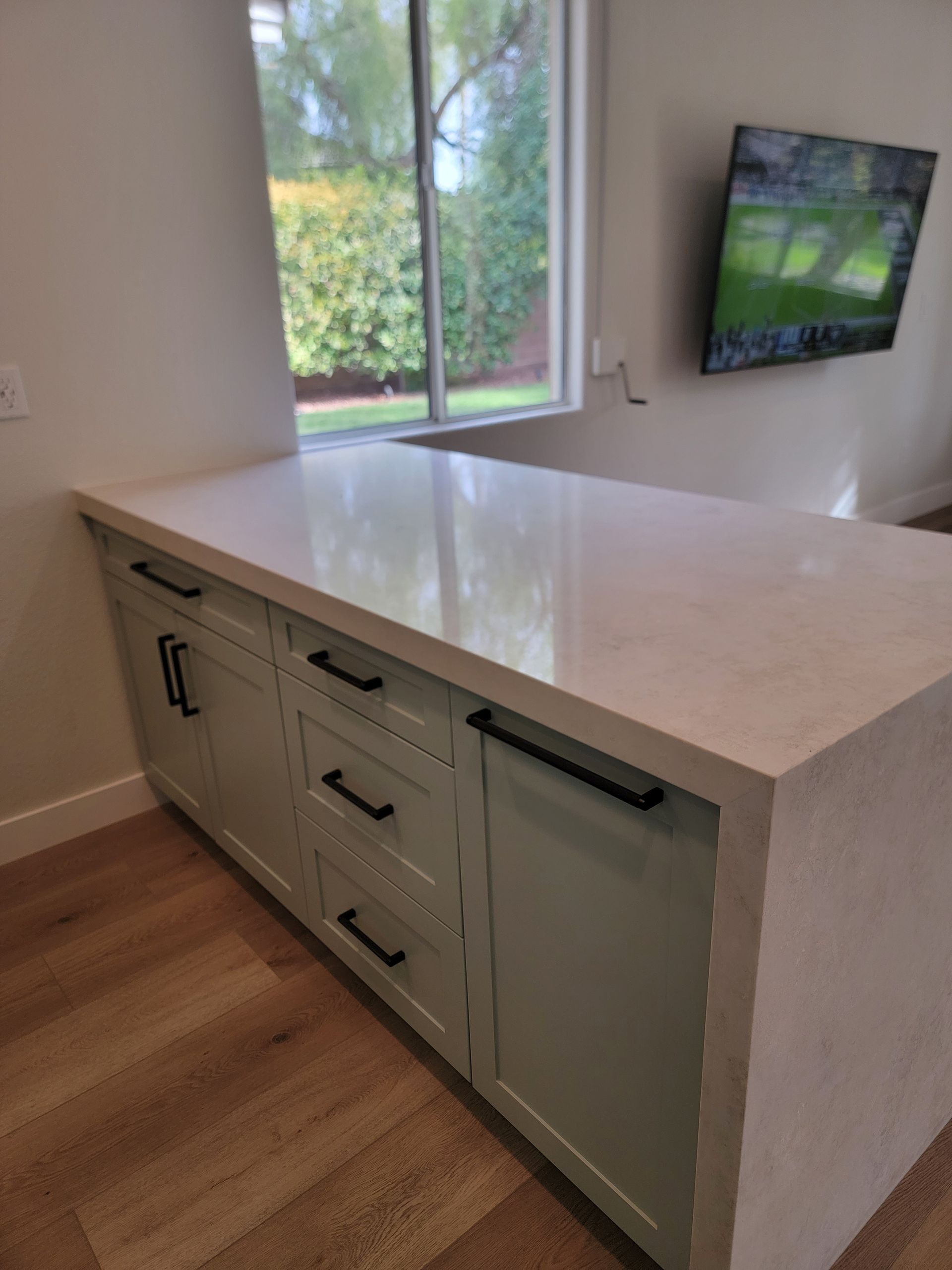 Kitchen island with light green cabinets, black handles, and light-colored countertop. TV and window in the background.