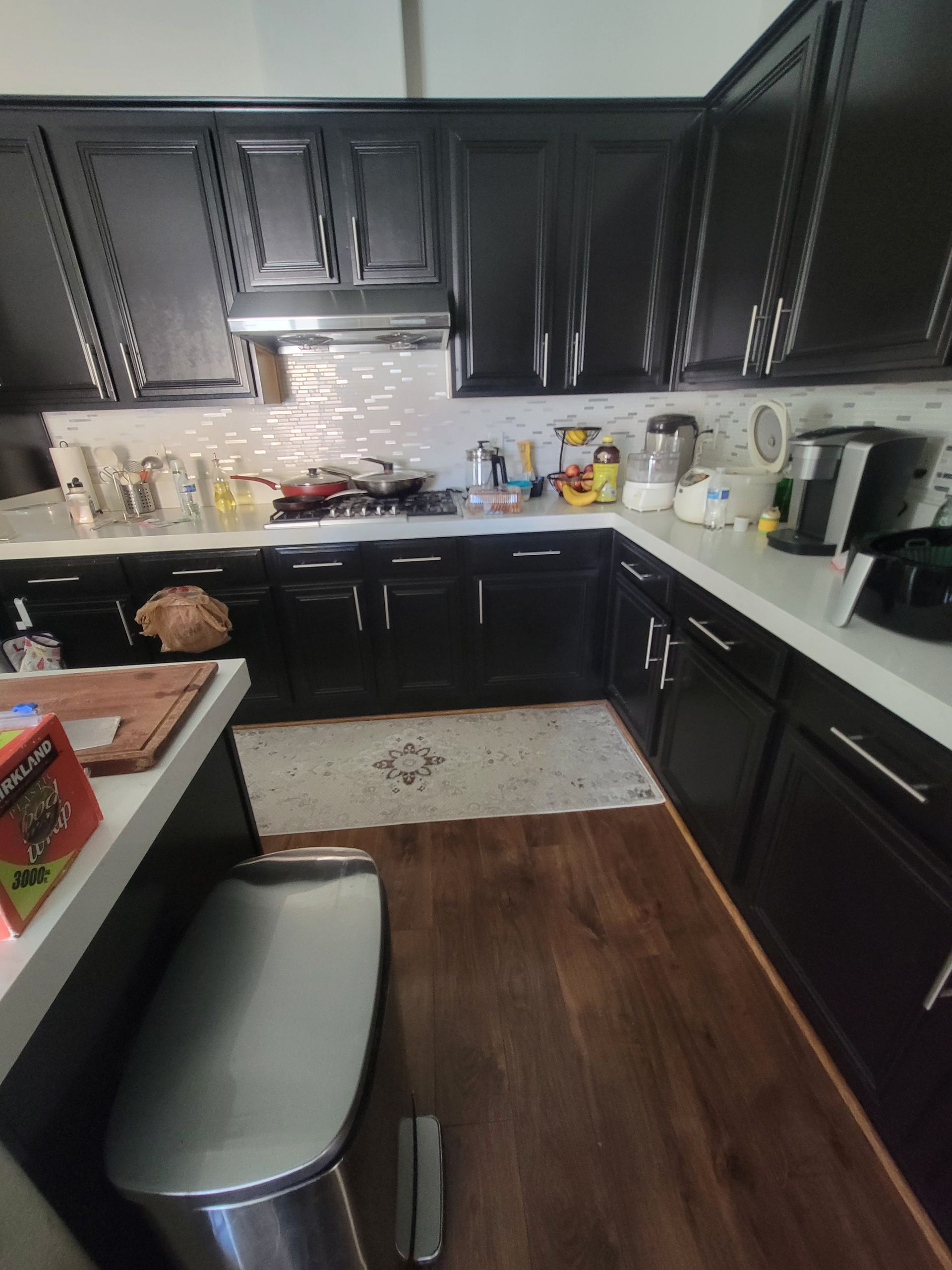 Black kitchen cabinets with white countertops, wooden floors, and a stainless steel trash can.