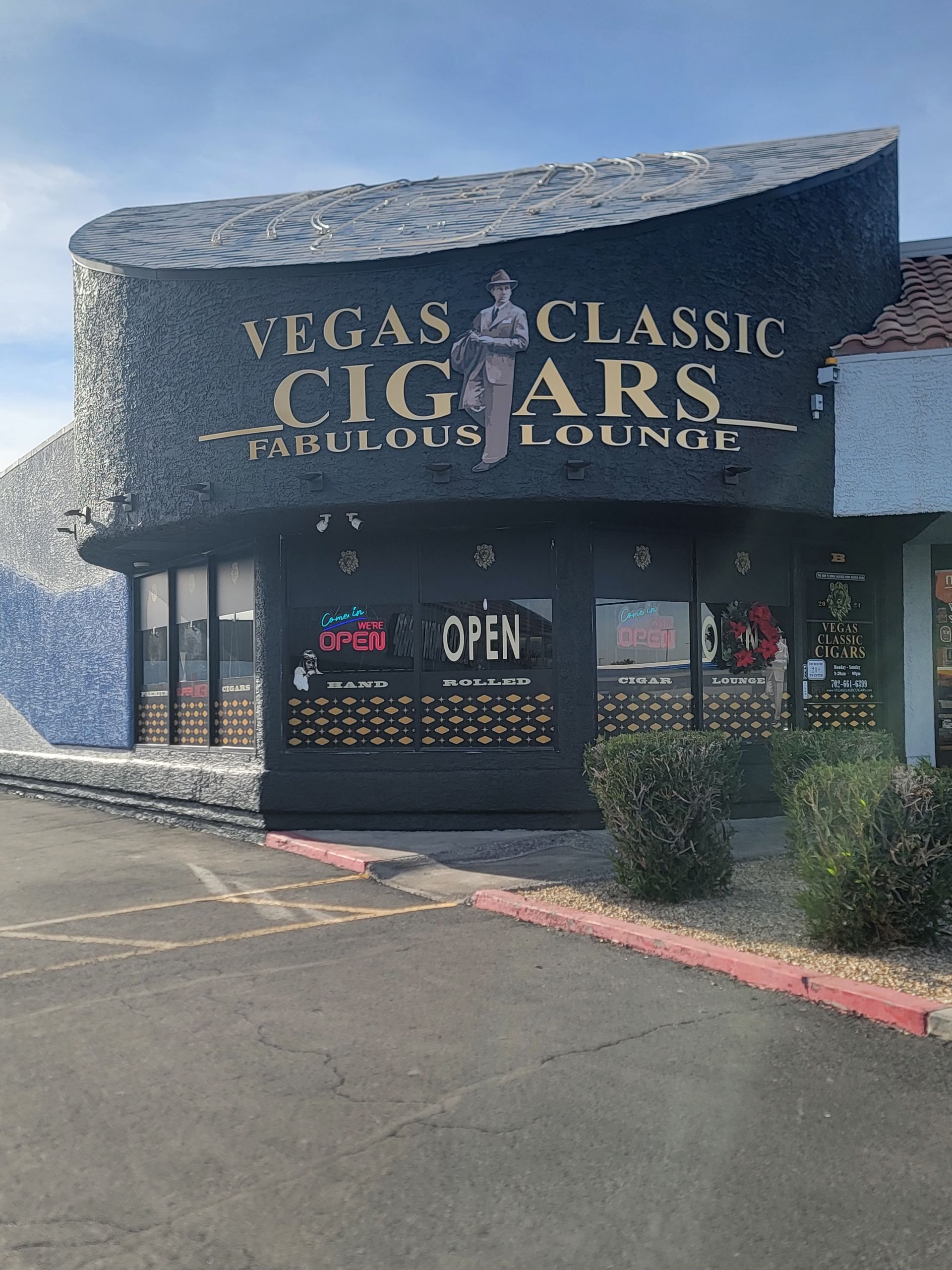 Vegas Classic Cigars, a brick building with sign and lounge. Open sign in window, bushes and parked car.
