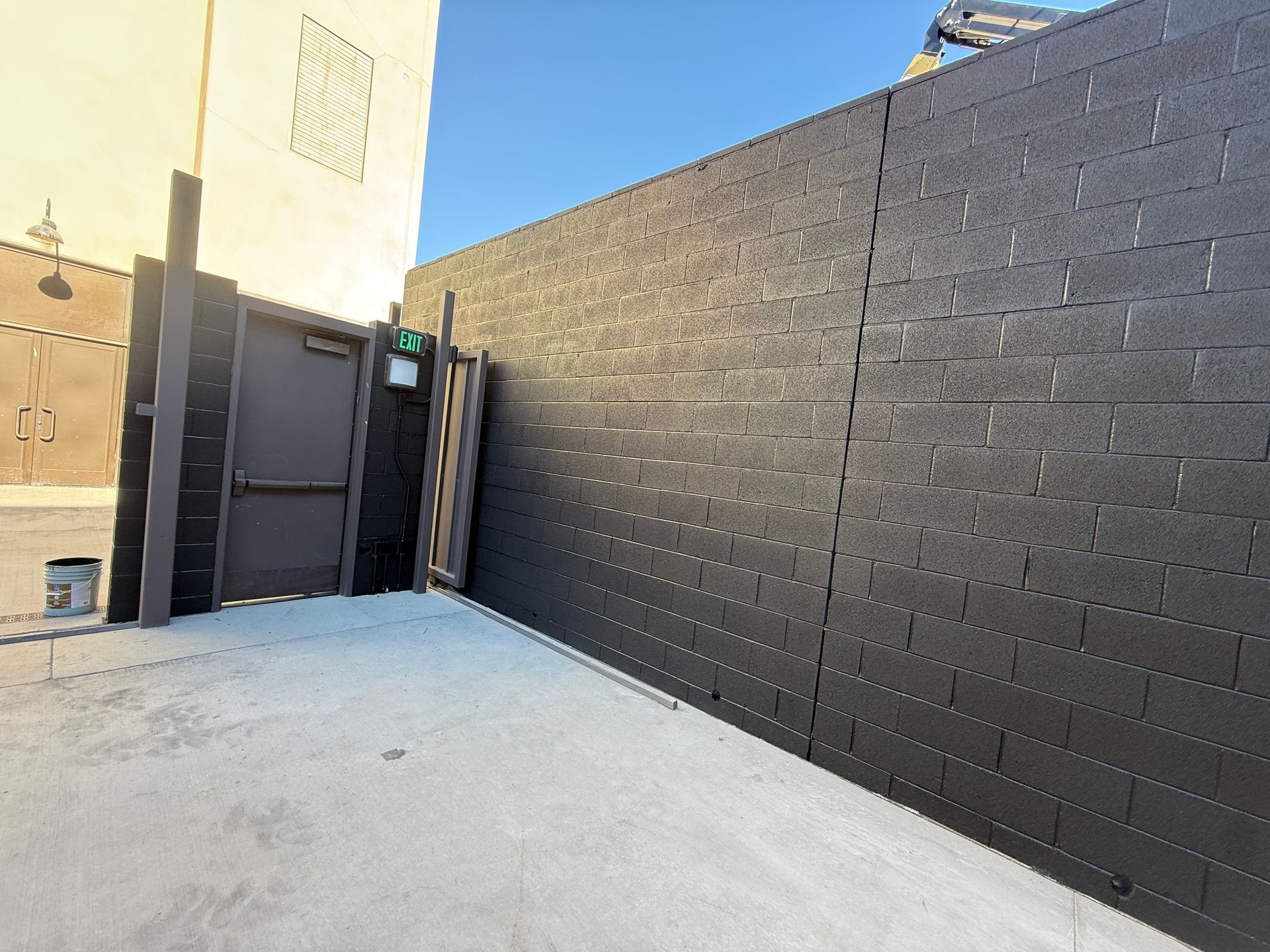Concrete alleyway with dark brick wall, door, and a glimpse of a building under a blue sky.