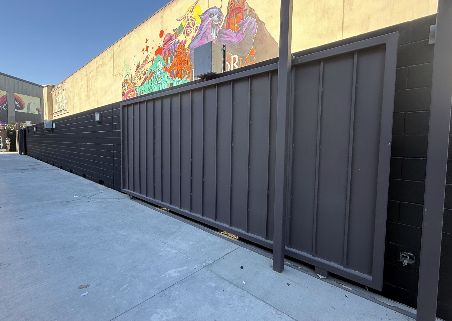Gray metal sliding gate on a city sidewalk, with a brick wall and mural in the background.