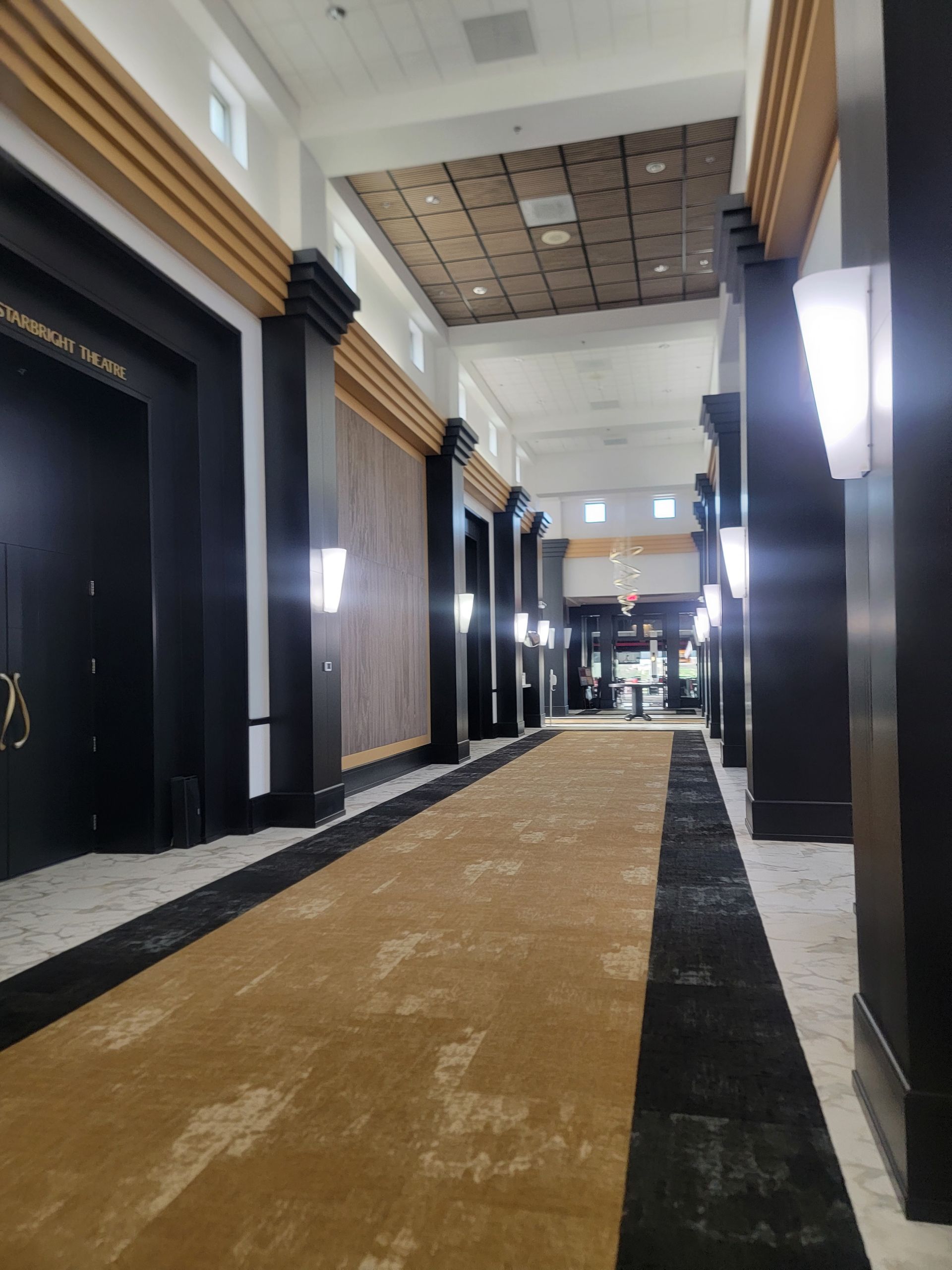 Long hallway with a gold and black carpet, black pillars, and sconce lighting.