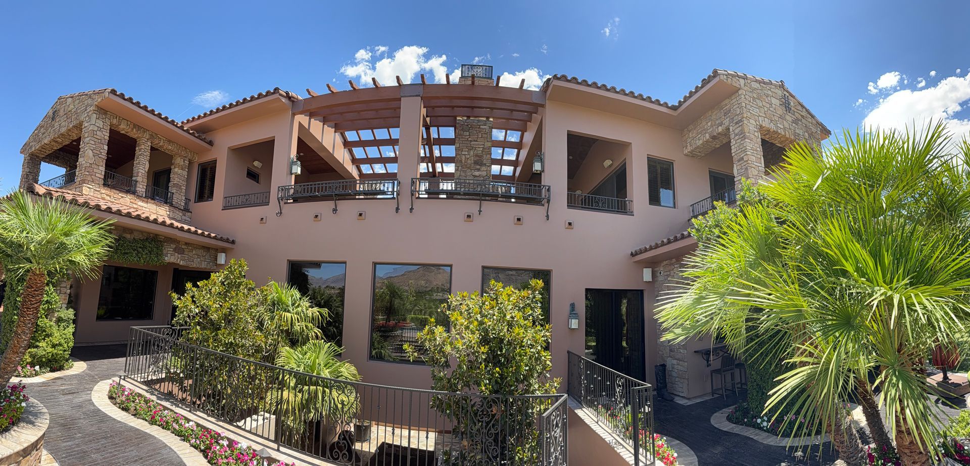 Large, light-colored building with dark windows and a wooden pergola, under a clear blue sky, surrounded by greenery.
