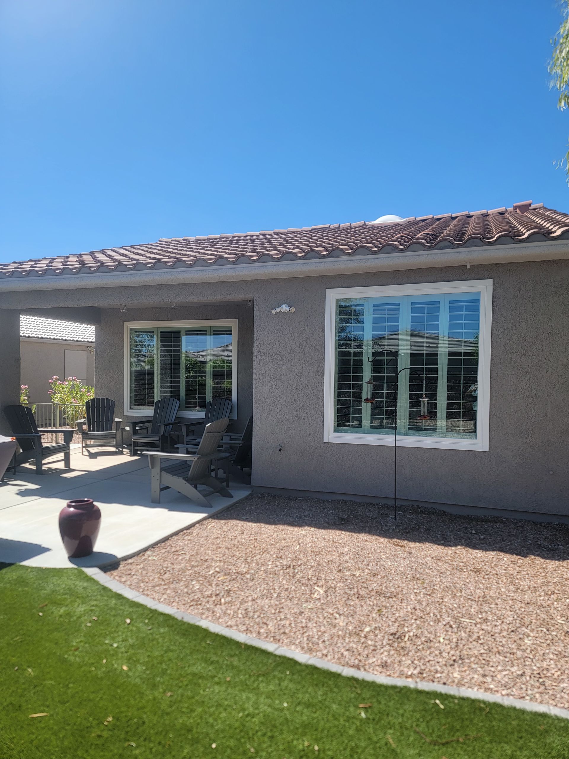 Backyard patio with gravel, artificial grass, and house with windows under a blue sky.
