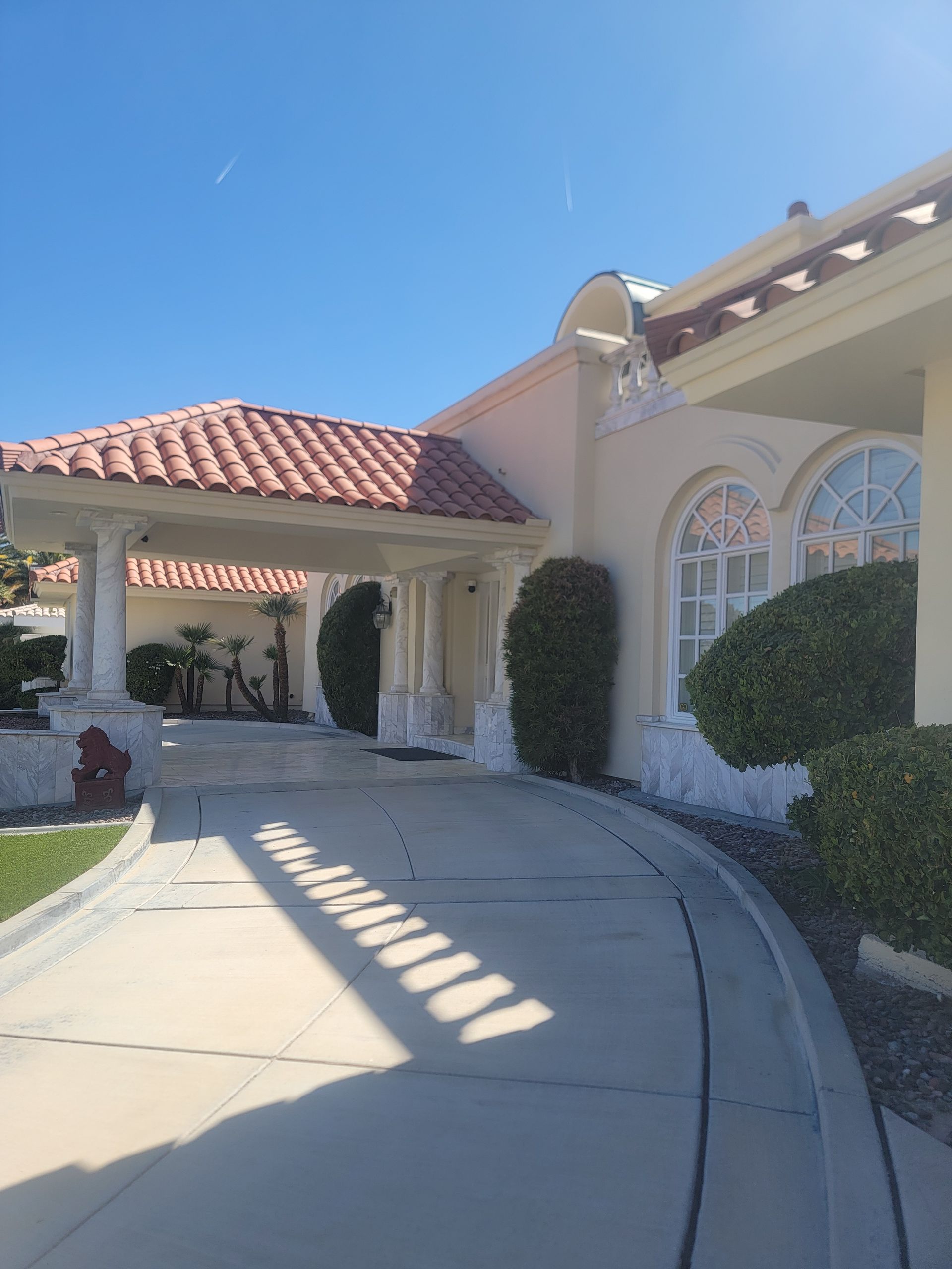 Beige stucco home with red tile roof, arched windows, and a curved driveway under a clear, blue sky.