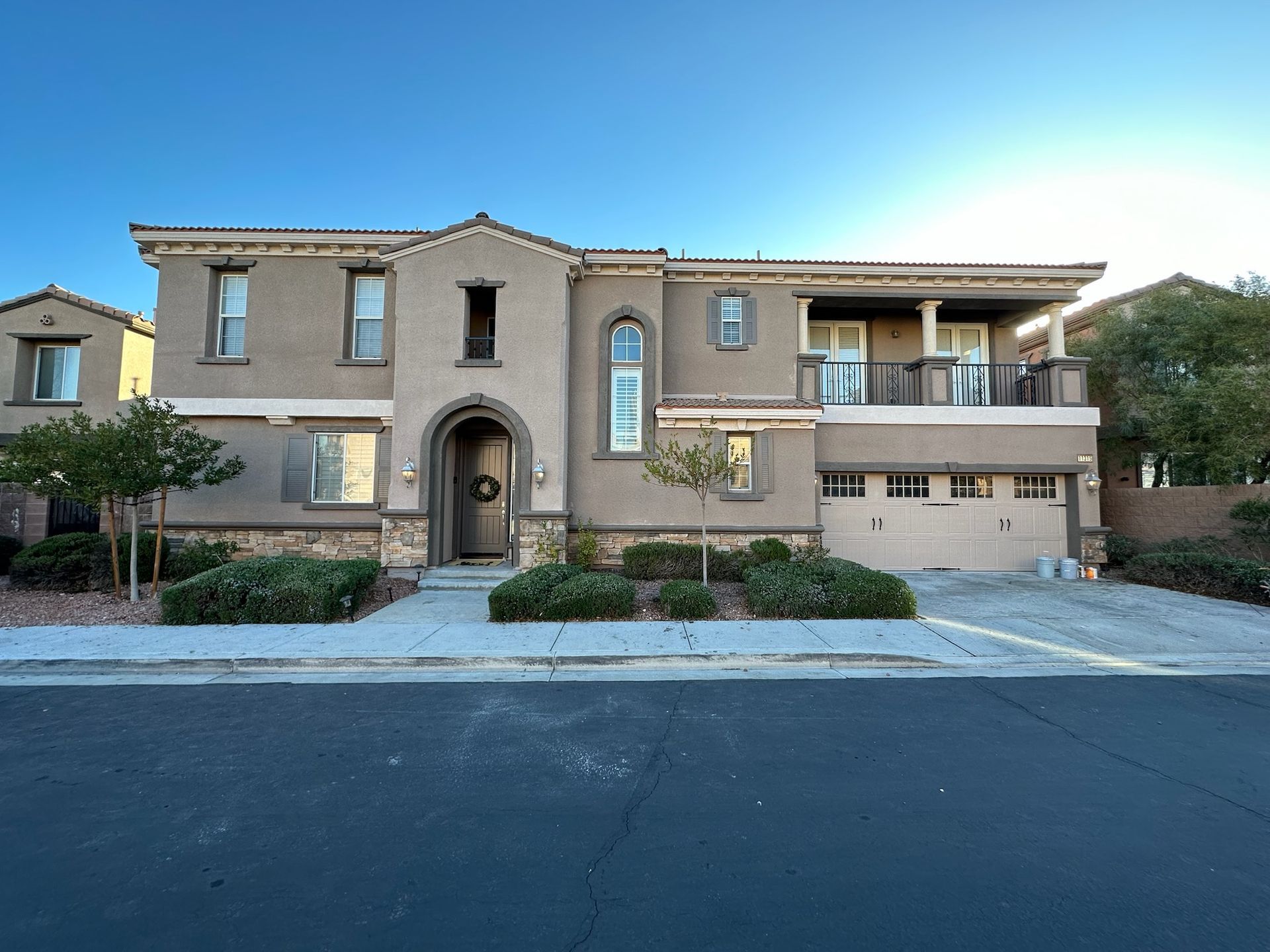 Two-story beige stucco house with a two-car garage, small trees, and bushes lining the front.