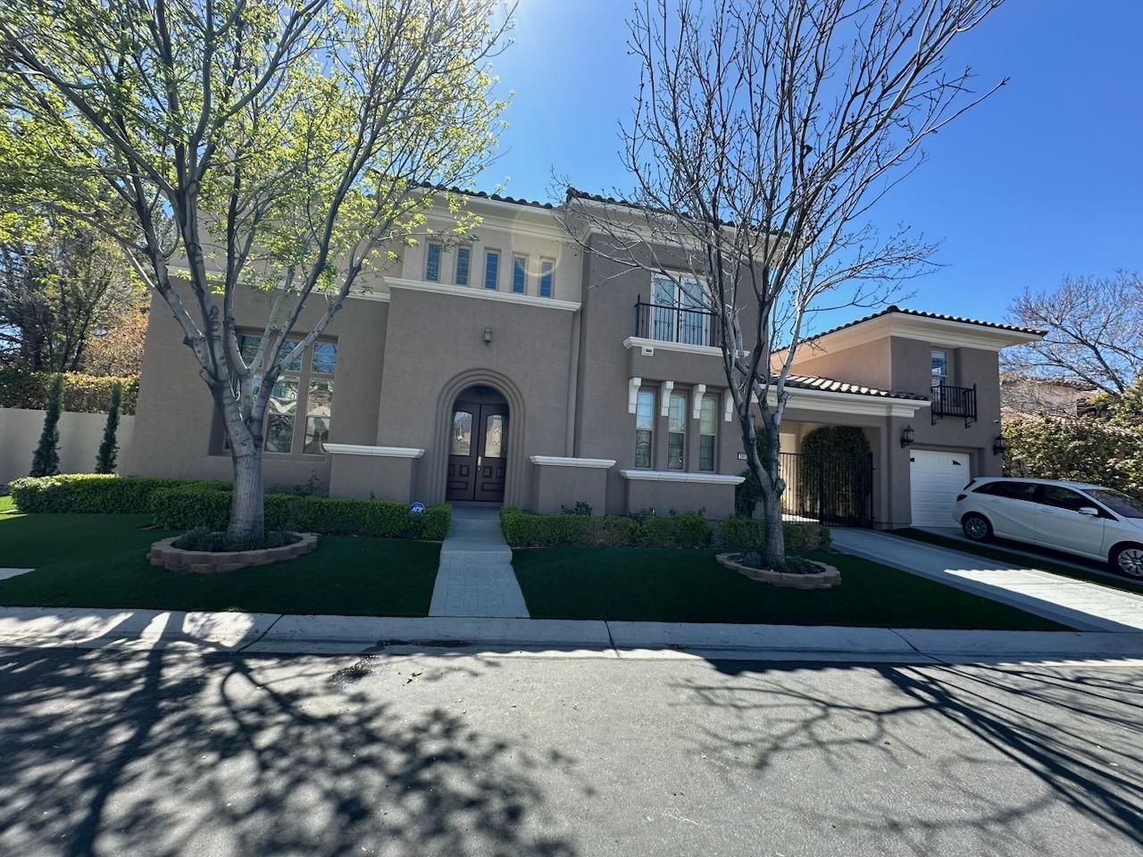 Two-story beige house with arched entry, two trees in front, green lawn, blue sky, and a car in the driveway.