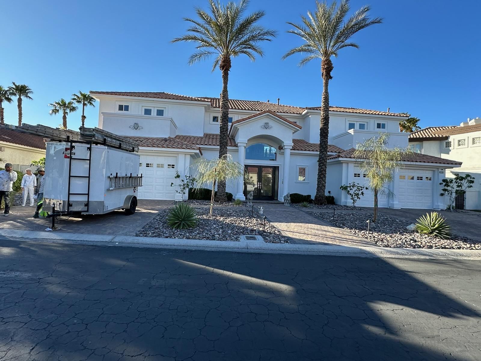 Large, two-story white house with palm trees, a trailer, and blue sky.