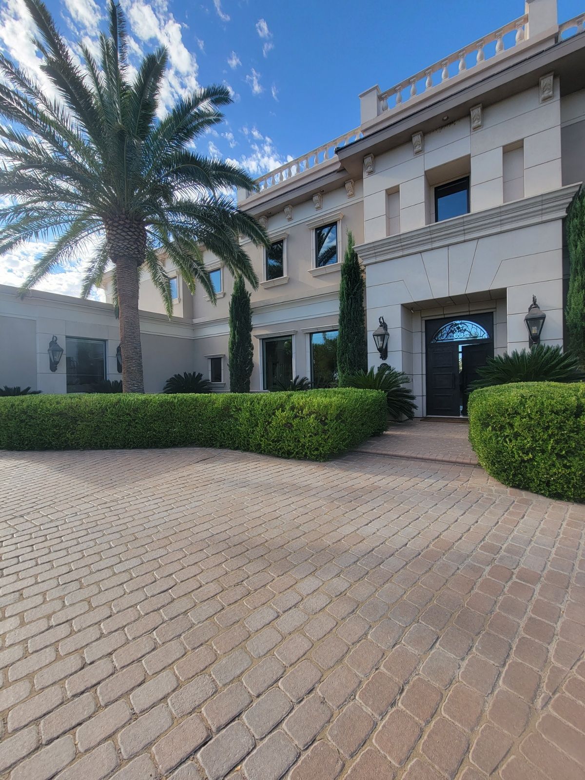 Exterior of a large beige house with a brick driveway, palm tree, and manicured hedges on a sunny day.