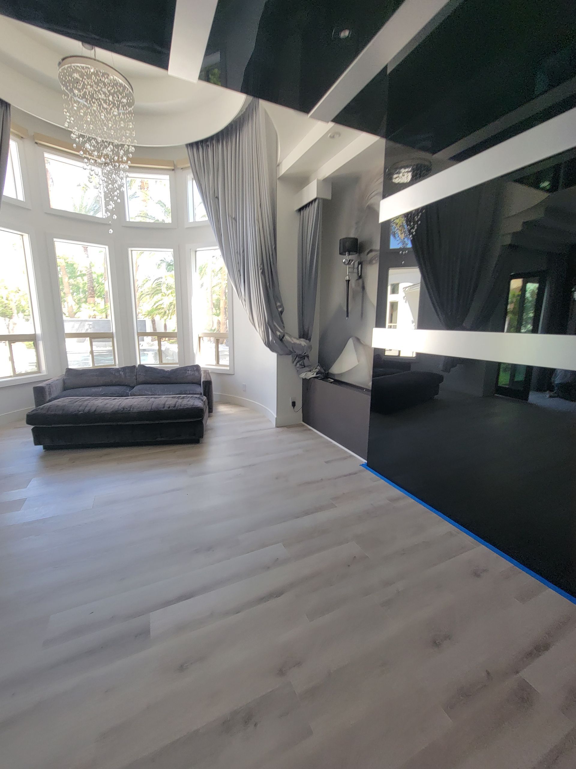 Living room with reflective black ceiling, grey couch, sheer curtains, and light wood-look flooring.
