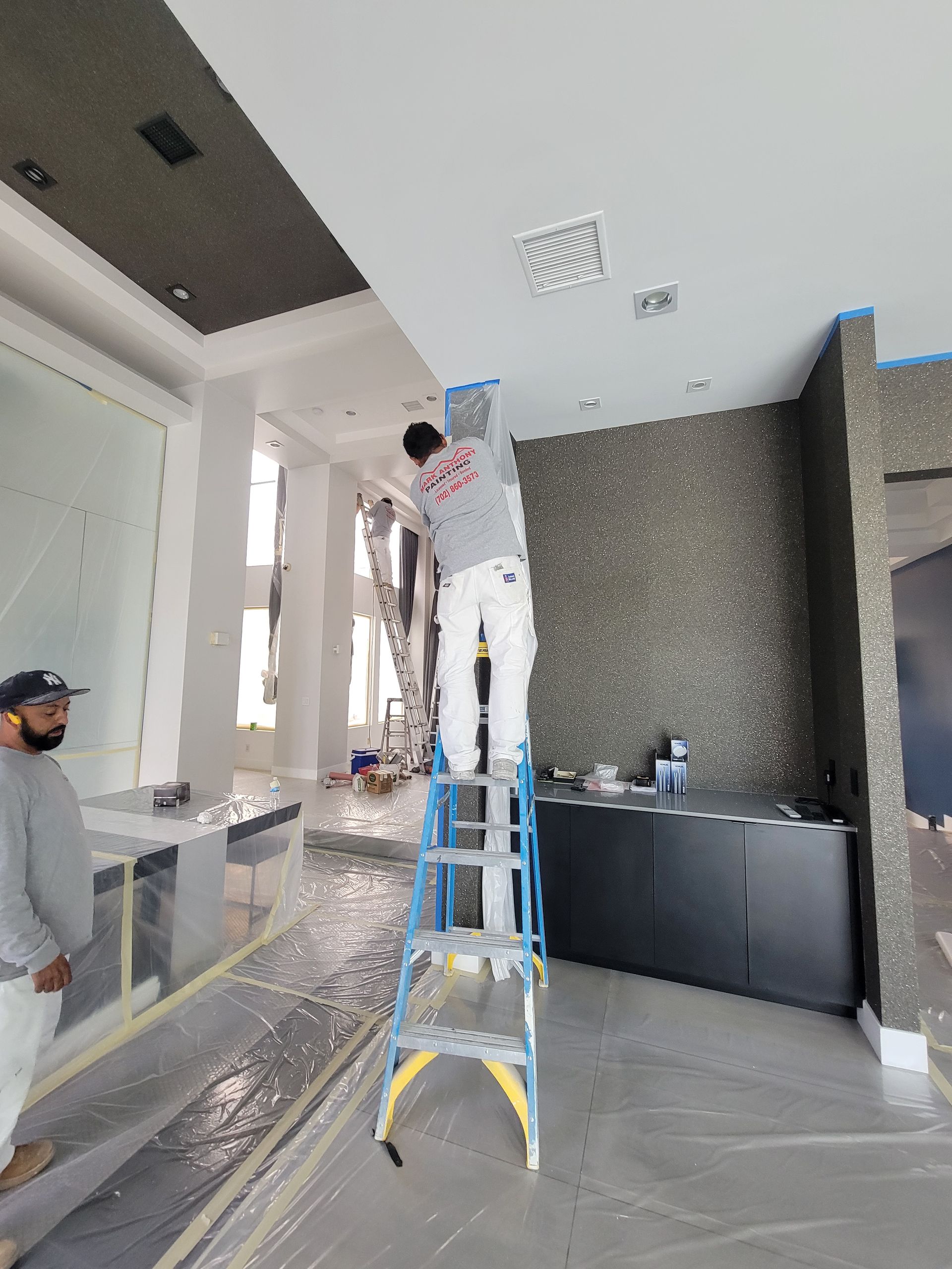 Two painters working in a room; one on a ladder painting a ceiling, another looking on. Room is under construction.