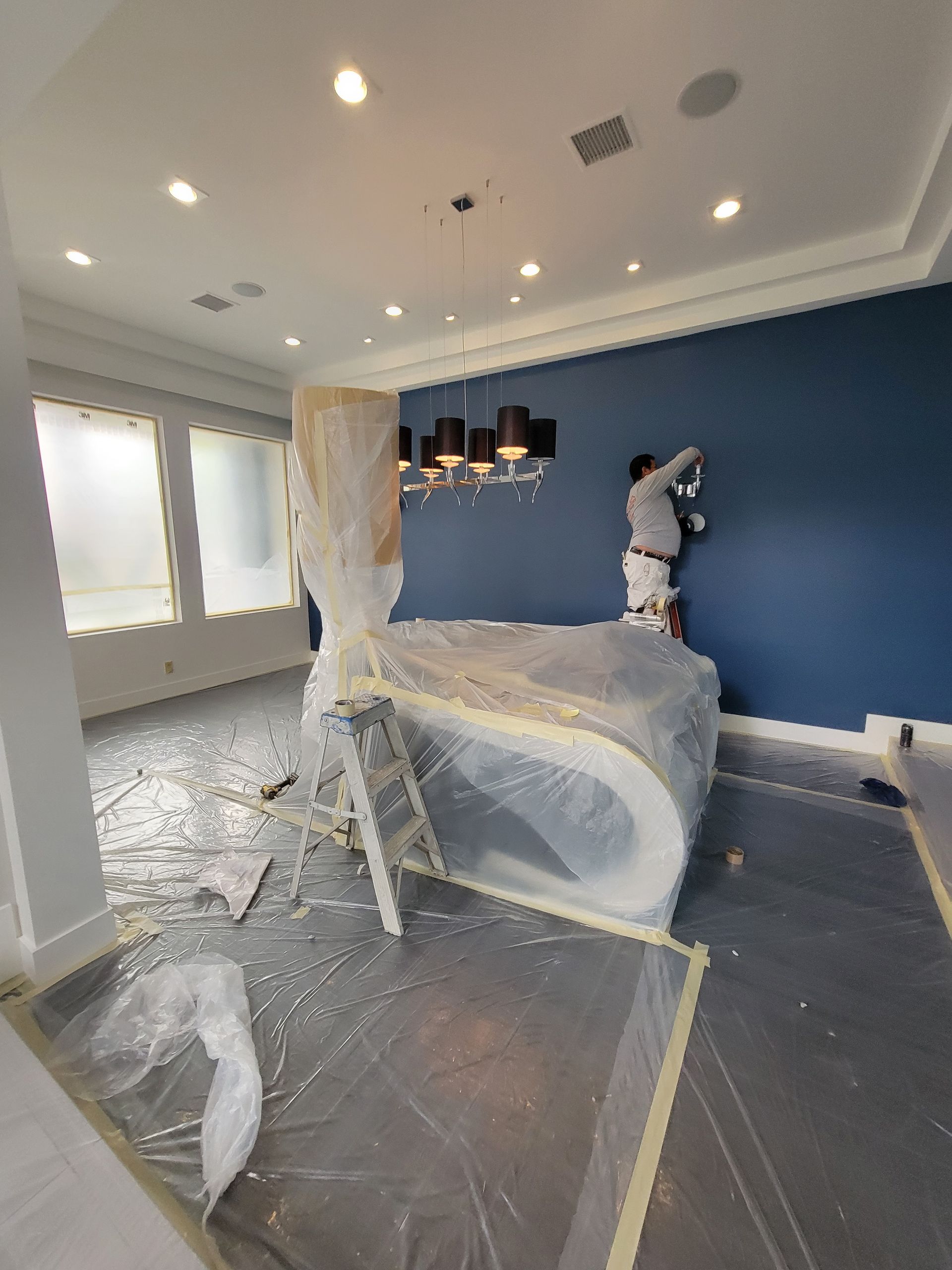Interior painting in progress: a painter on a ladder applying blue paint to a wall, with a covered counter and floor.