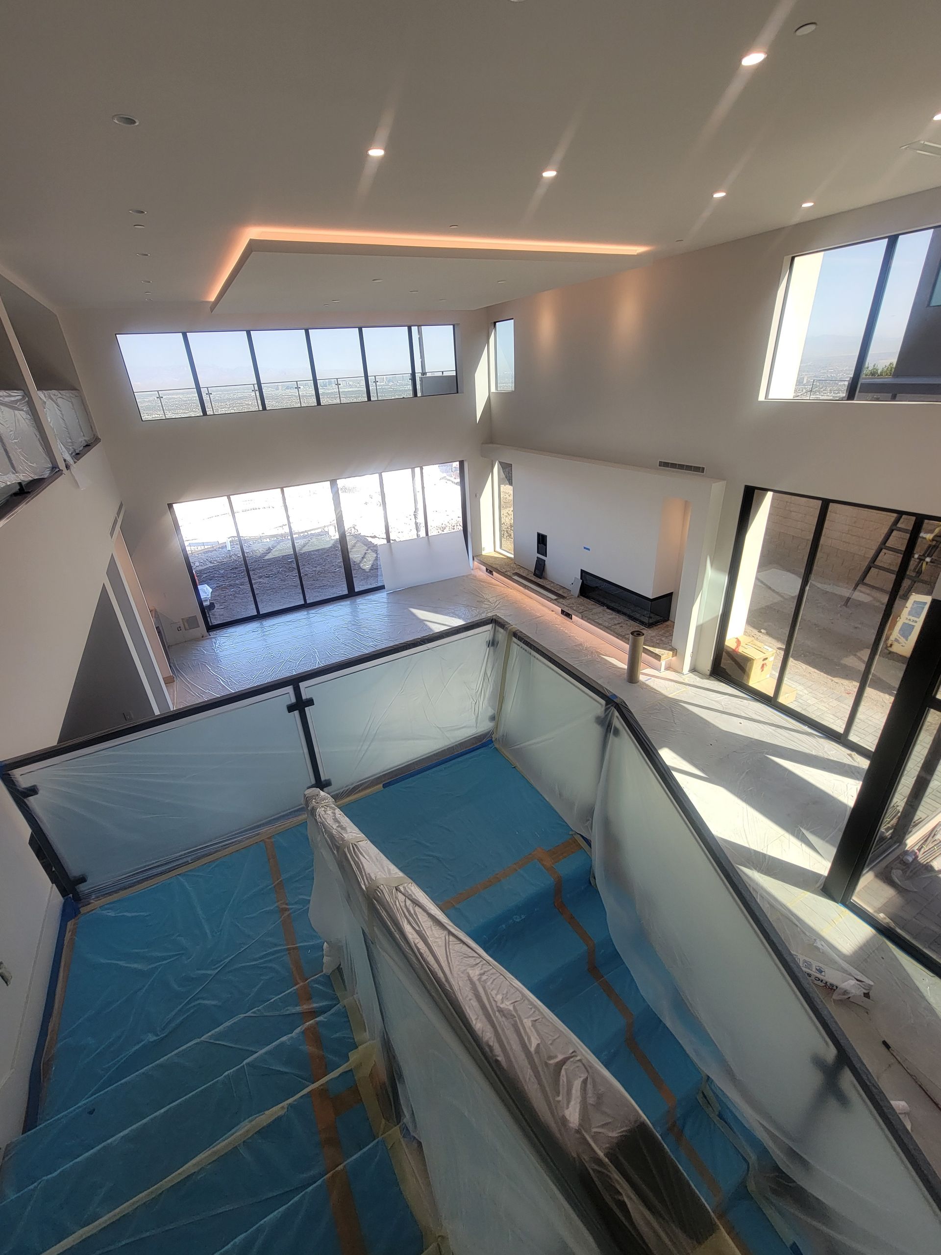 Modern, multi-story interior with glass railings and large windows; light blue tarp covers a pool or similar feature.