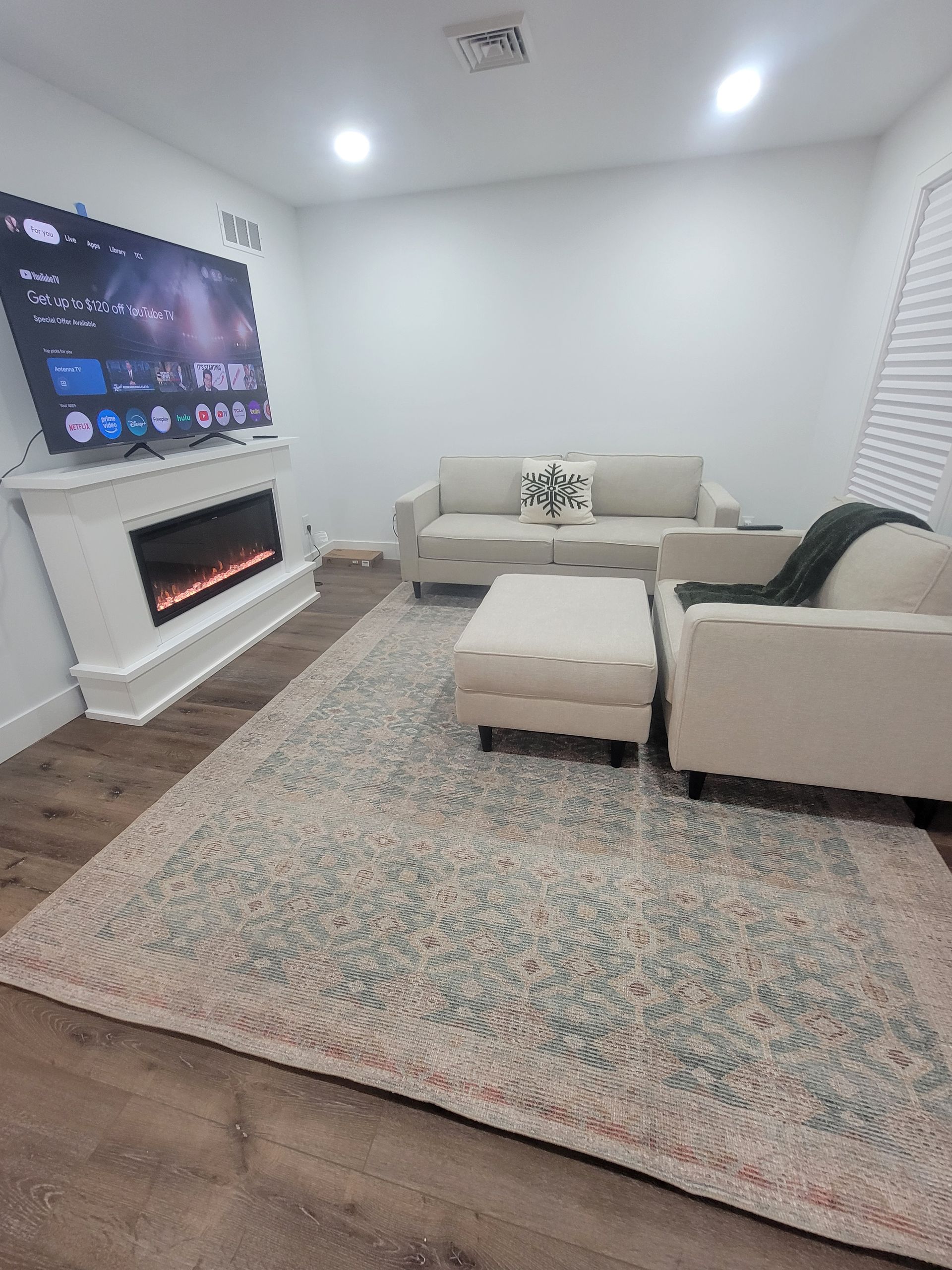 Living room with a white sofa, armchair, and ottoman on a patterned rug; a fireplace with a TV above.