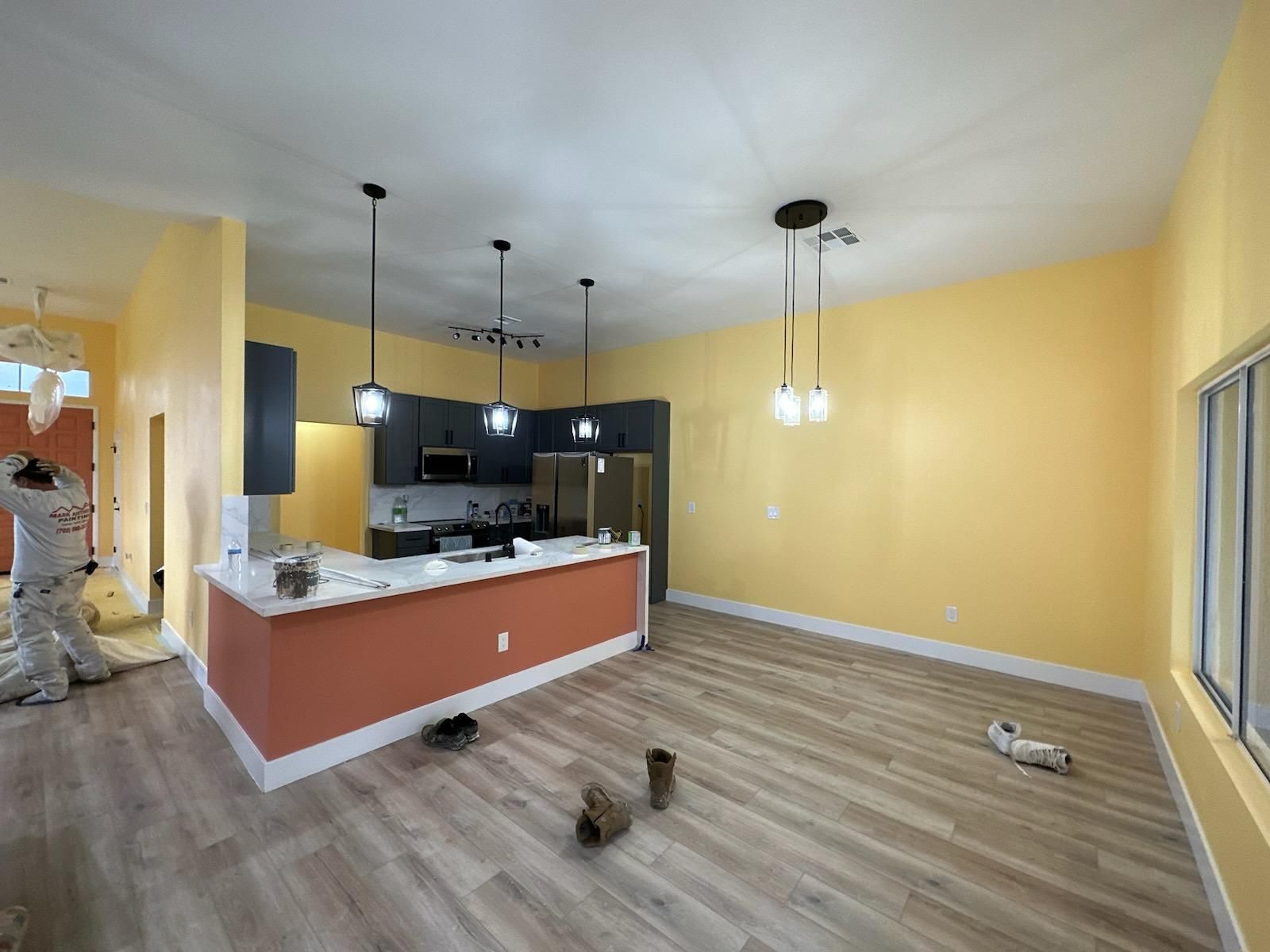 A room with yellow walls, a kitchen with black cabinets and coral island, wooden floors, and hanging lights.