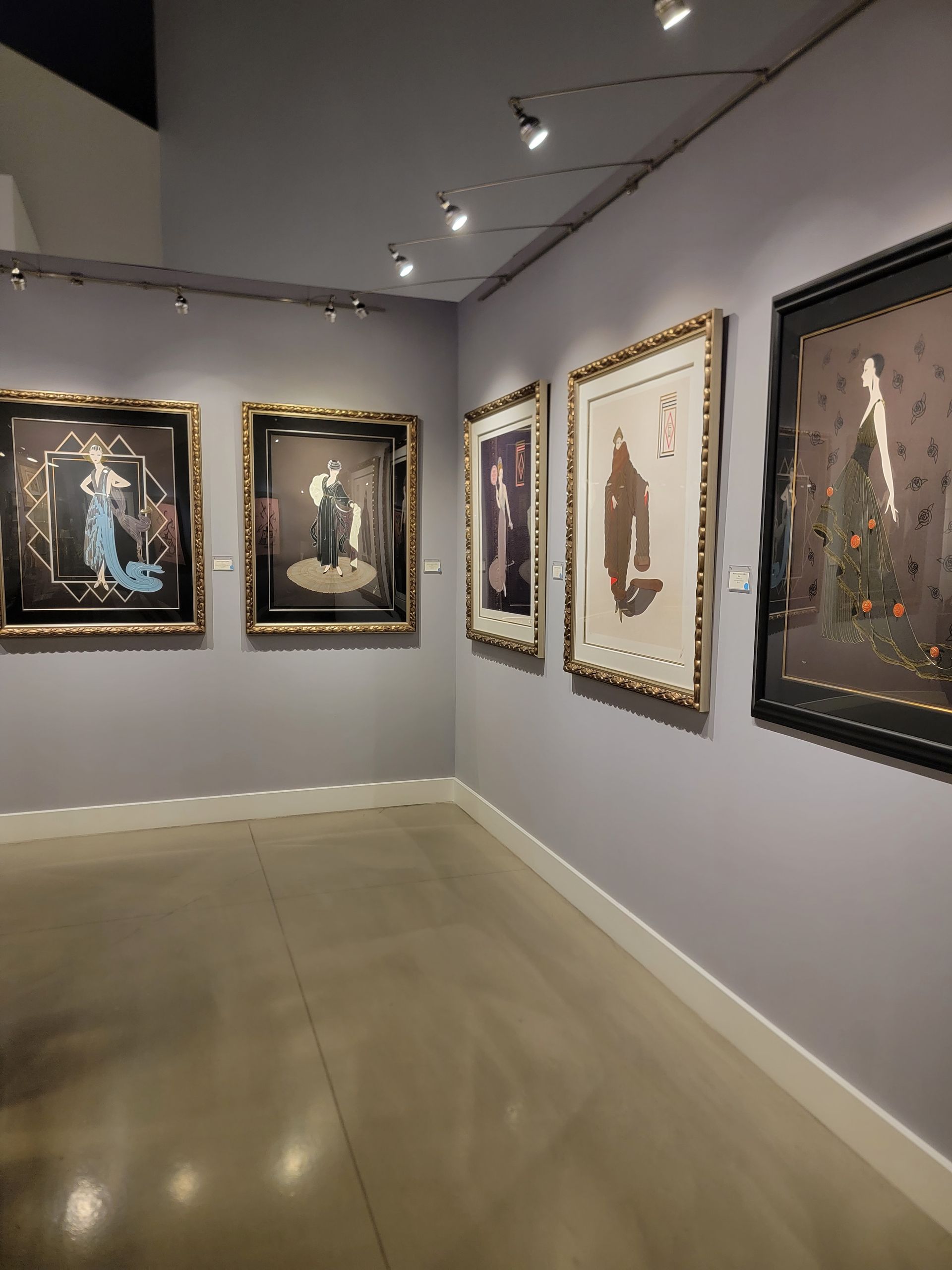 Art gallery corner with framed pictures on grey walls. Track lighting illuminates the artwork, and the floor is shiny.