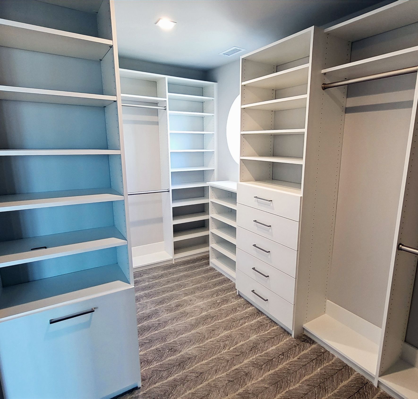 White custom closet with shelves, drawers, and hanging rods. Dark wood-look flooring and a round window.