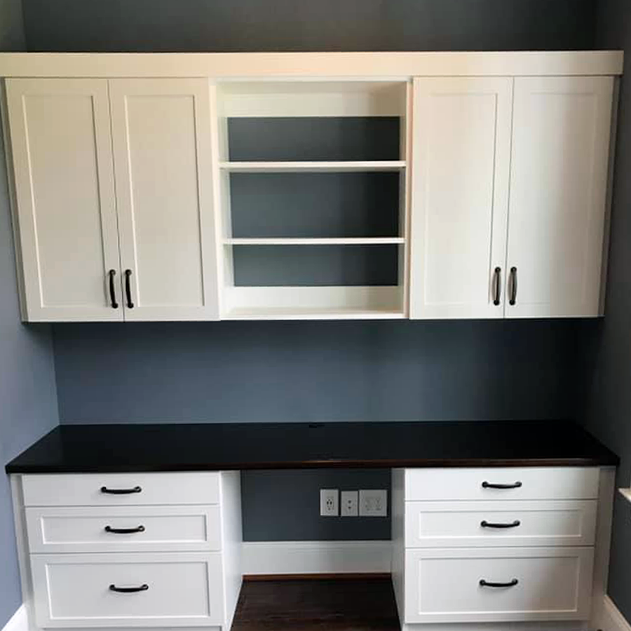 Built-in white desk with upper cabinets and open shelves on a blue-gray wall. Black countertop and dark wood floor.