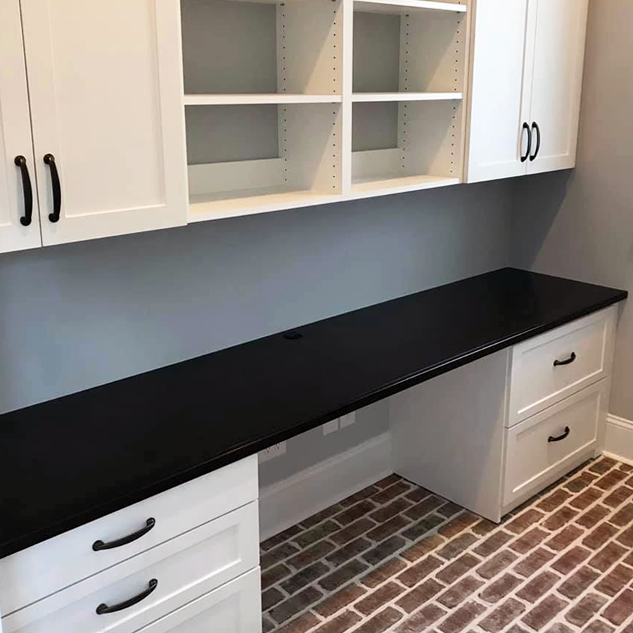 White home office built-in desk with black countertop and overhead storage; brick floor.