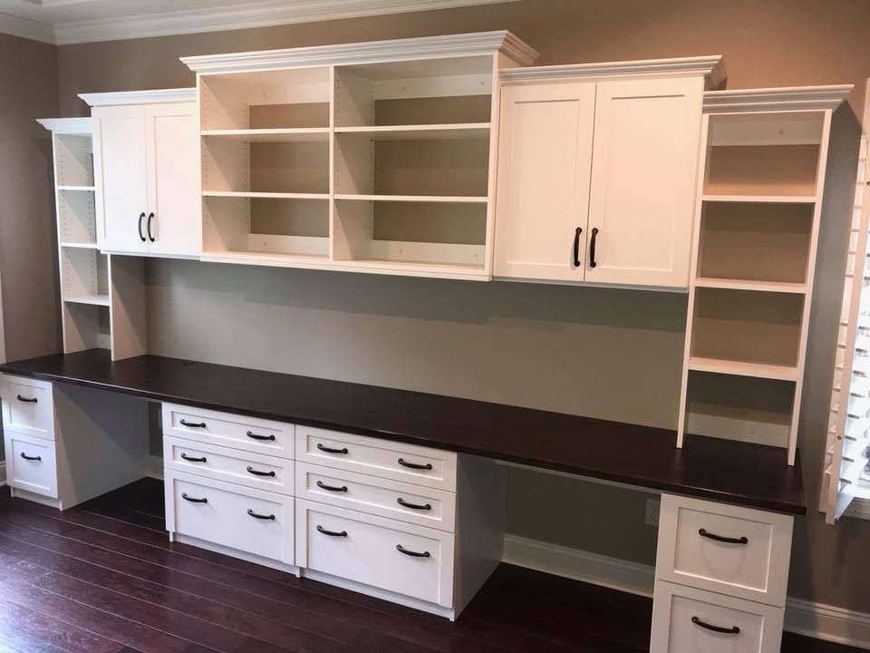 Built-in white desk with cabinets, shelves, and dark countertop against a light-colored wall.