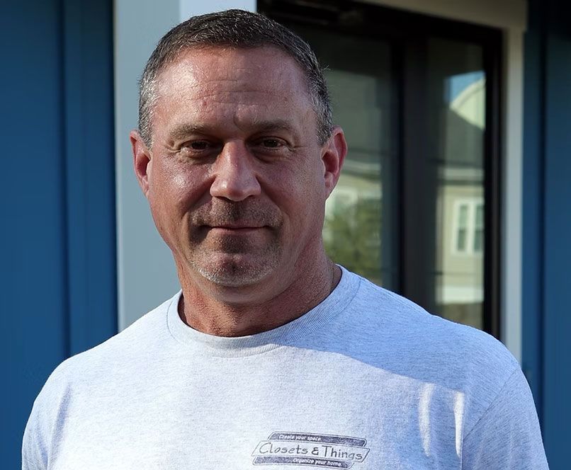Man in gray shirt smiles, stands outside a blue building.