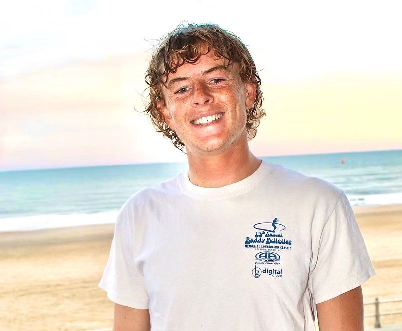 Man smiling in front of a beach. He wears a white shirt with a surfing logo and has curly hair.