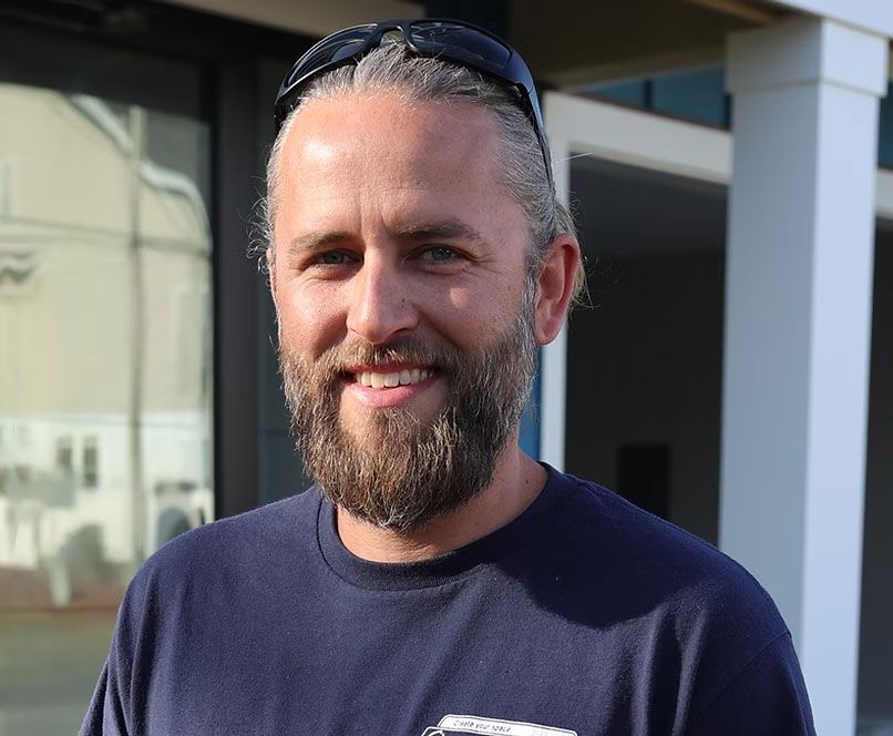 Man with graying hair and beard smiles outdoors, sunglasses on head, navy blue shirt.