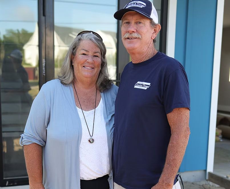Couple standing in front of a blue building with dark trim; woman in light blue cardigan, man in navy shirt and hat.