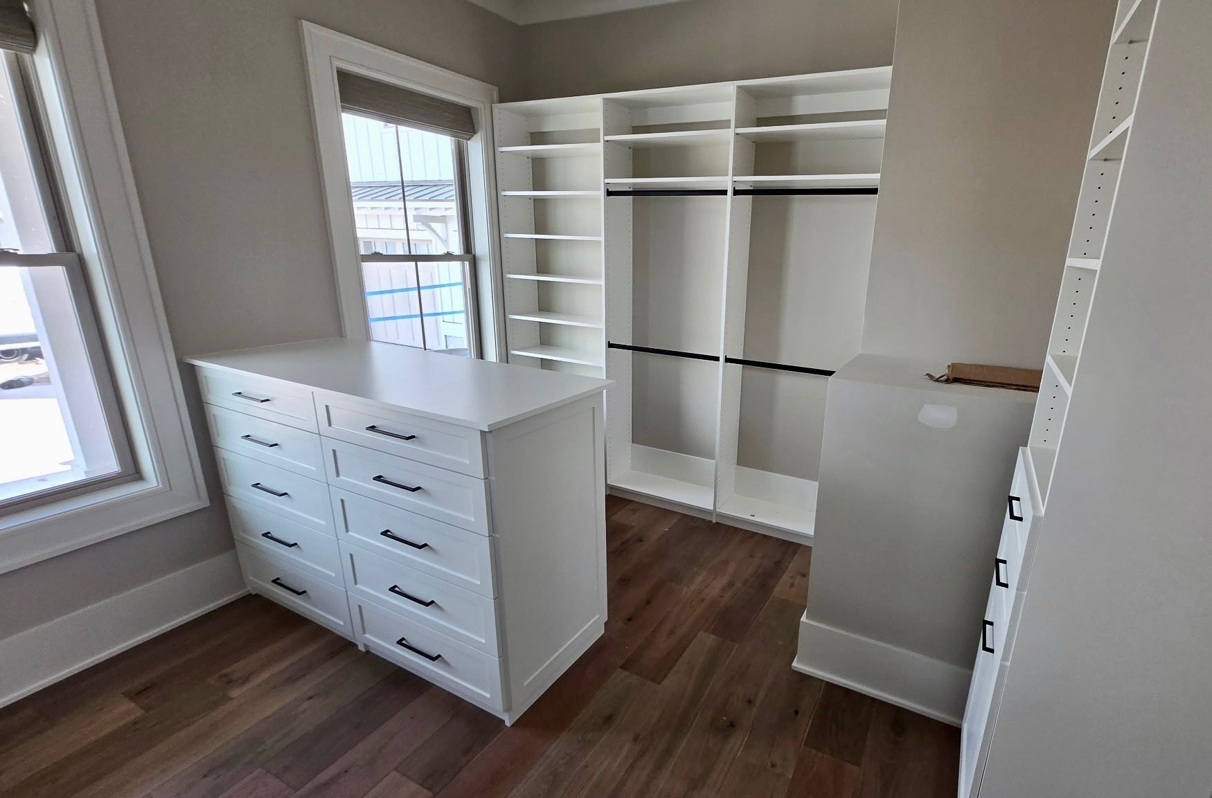 Empty walk-in closet with white shelves, drawers, and hanging rods on dark wood flooring.