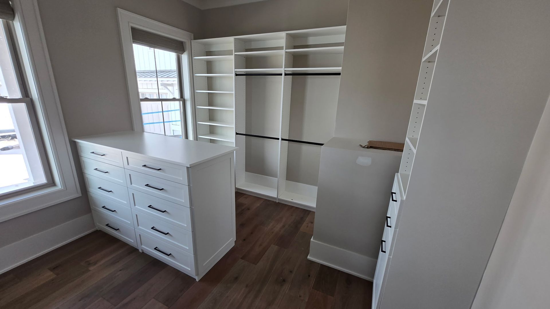 Empty walk-in closet with white shelving, drawers, and brown wood flooring beside a window.