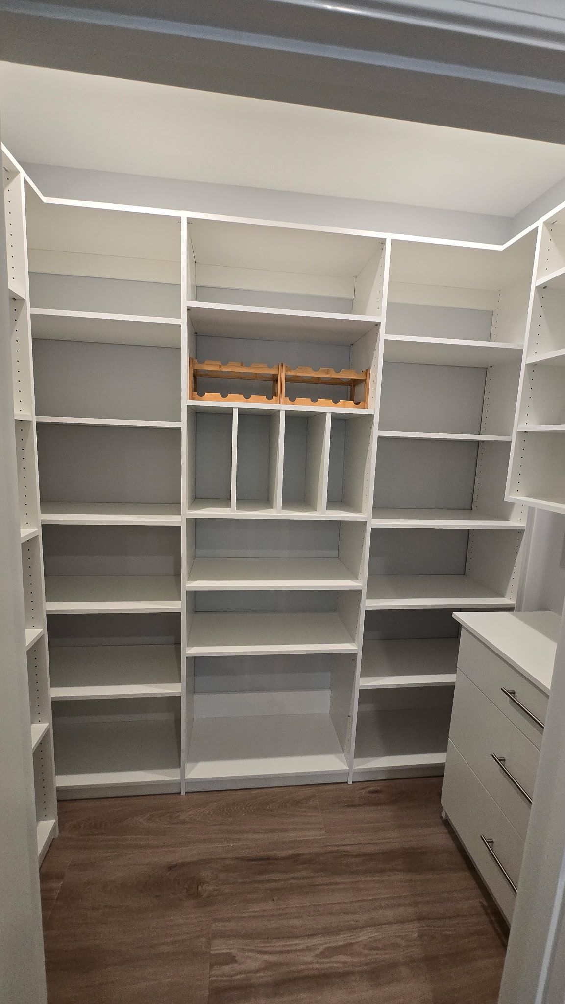 Empty walk-in closet with white shelving and wood flooring, featuring built-in drawers on the right.