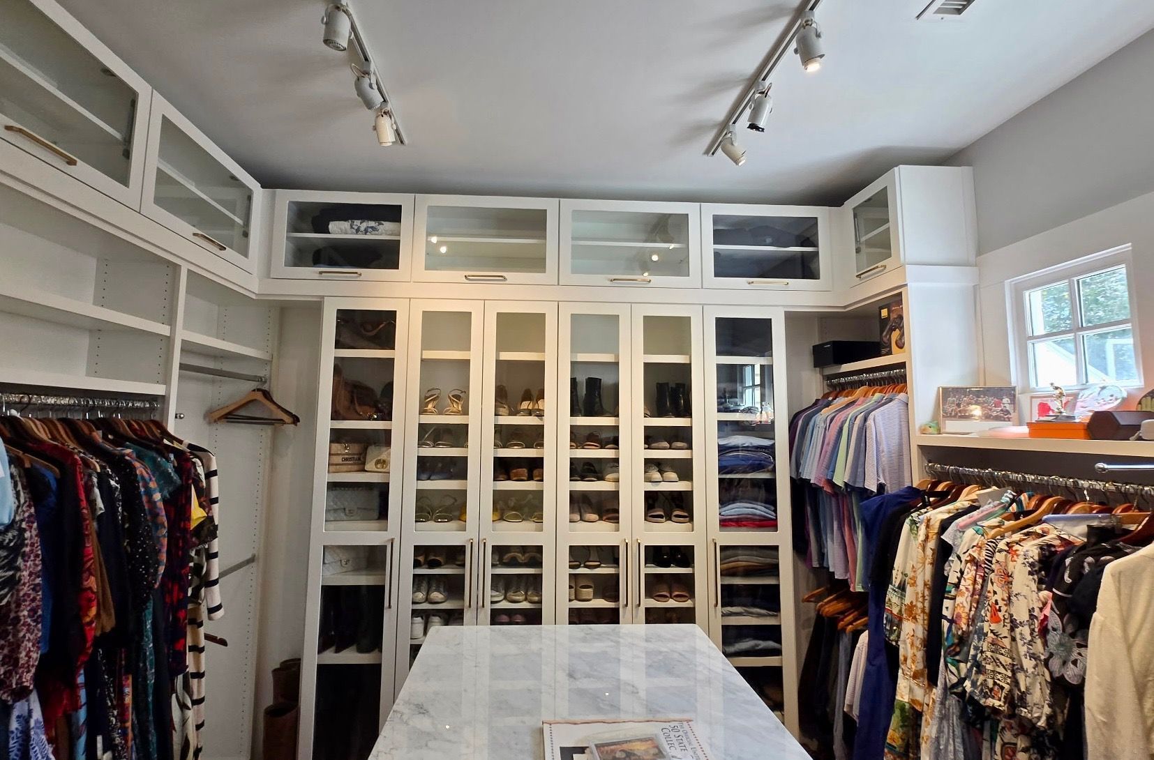 Bright walk-in closet with white shelving, hanging clothes, and a glass-topped island table