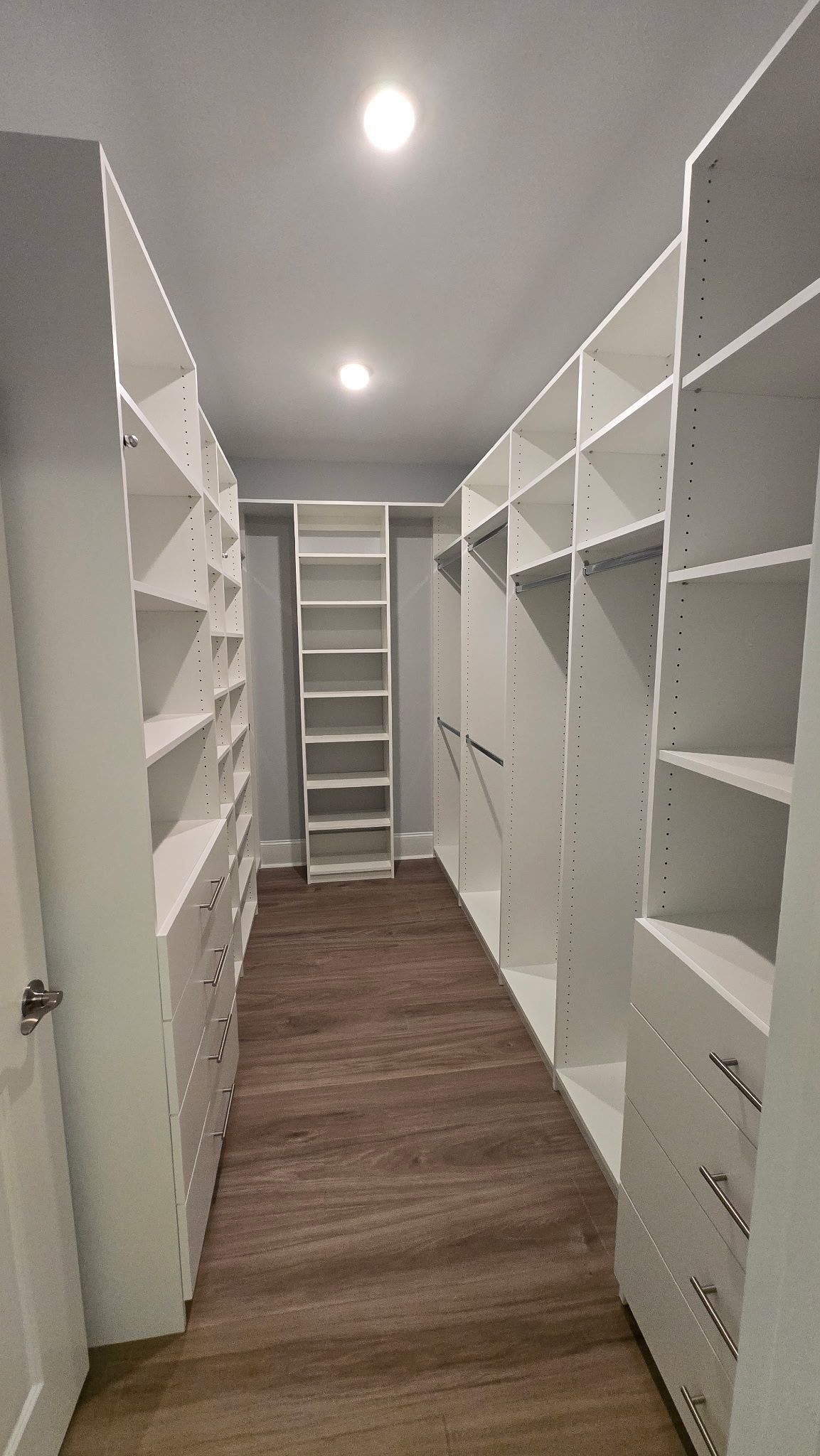Narrow walk-in closet with white shelving on both sides and wood-look flooring, lit by a ceiling light.