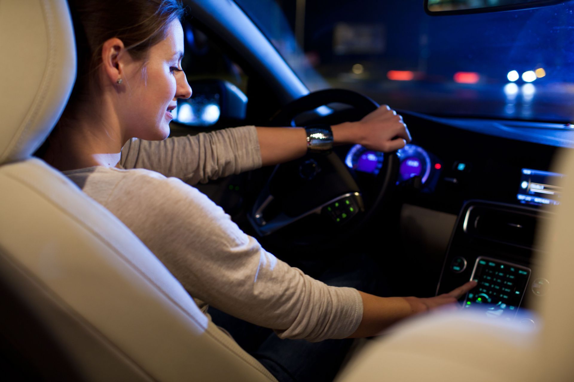 Woman driving a car at night, smiling and adjusting the dashboard controls. Interior lit in blue.