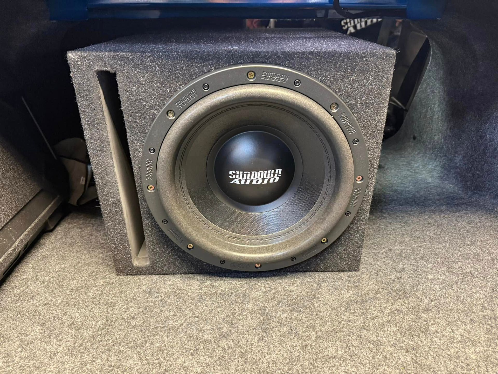 Subwoofer in a car trunk, black speaker with gray enclosure, slot port on left side, mounted on gray carpet.