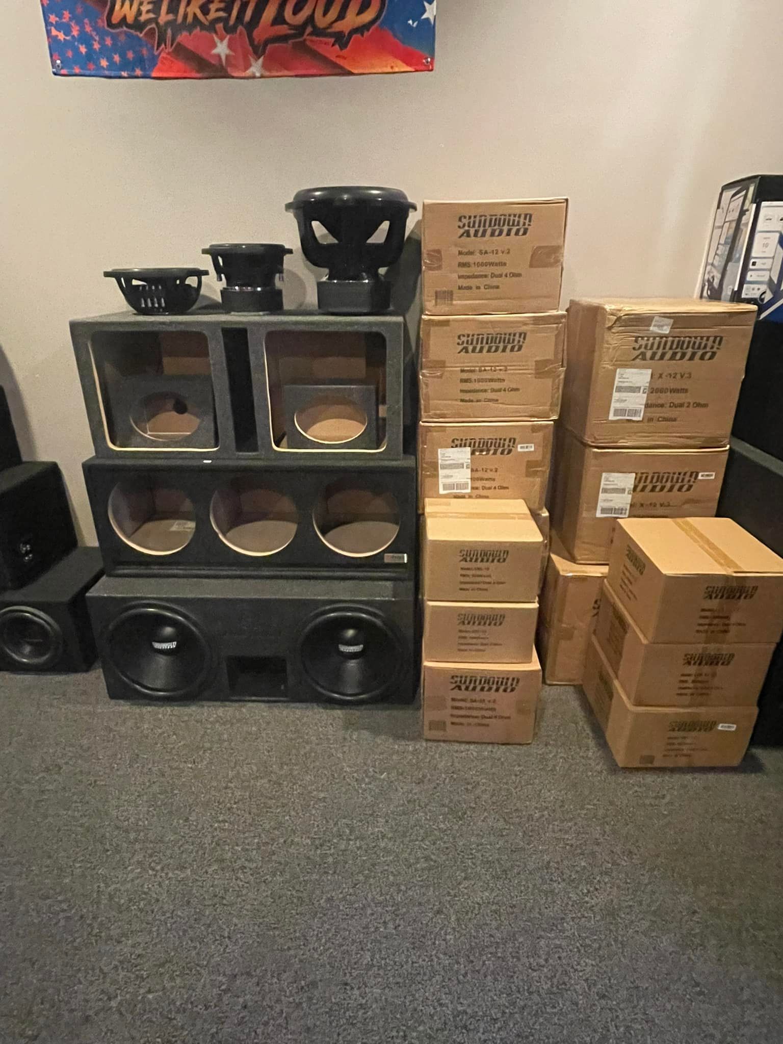 A collection of subwoofer boxes, speakers, and speaker components are stacked against a wall with cardboard boxes.