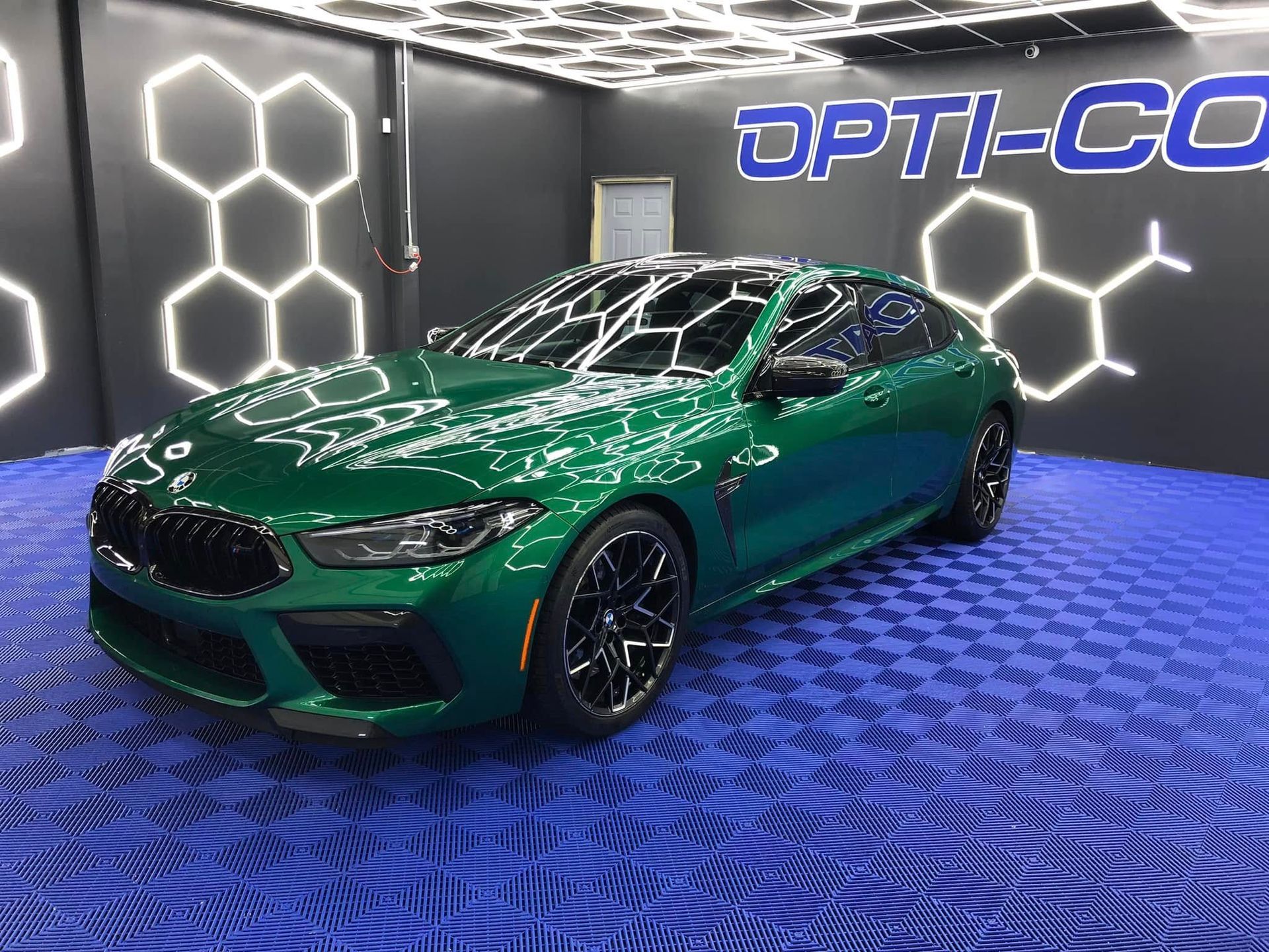 Green BMW M8 Gran Coupe in a detailing studio, lit by honeycomb lights and blue flooring.