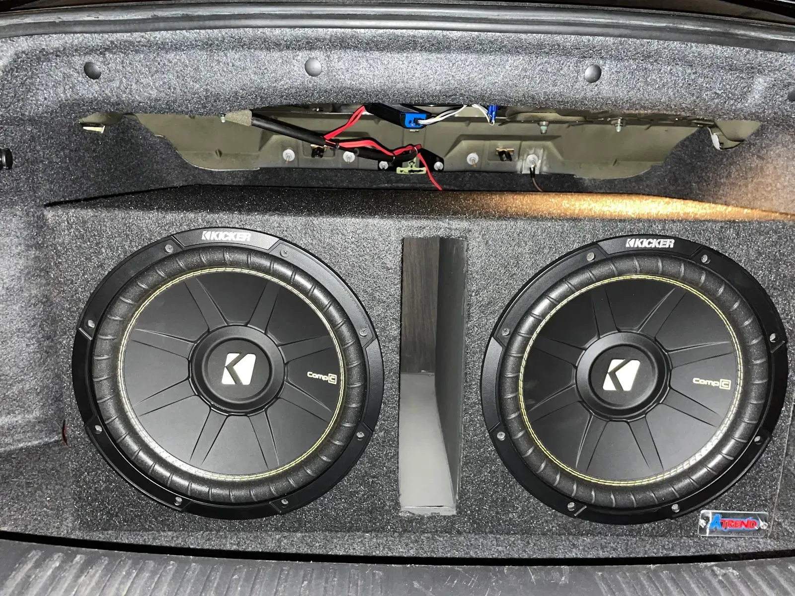 Two Kicker subwoofers mounted in a car trunk. Gray textured enclosure. Wiring visible.